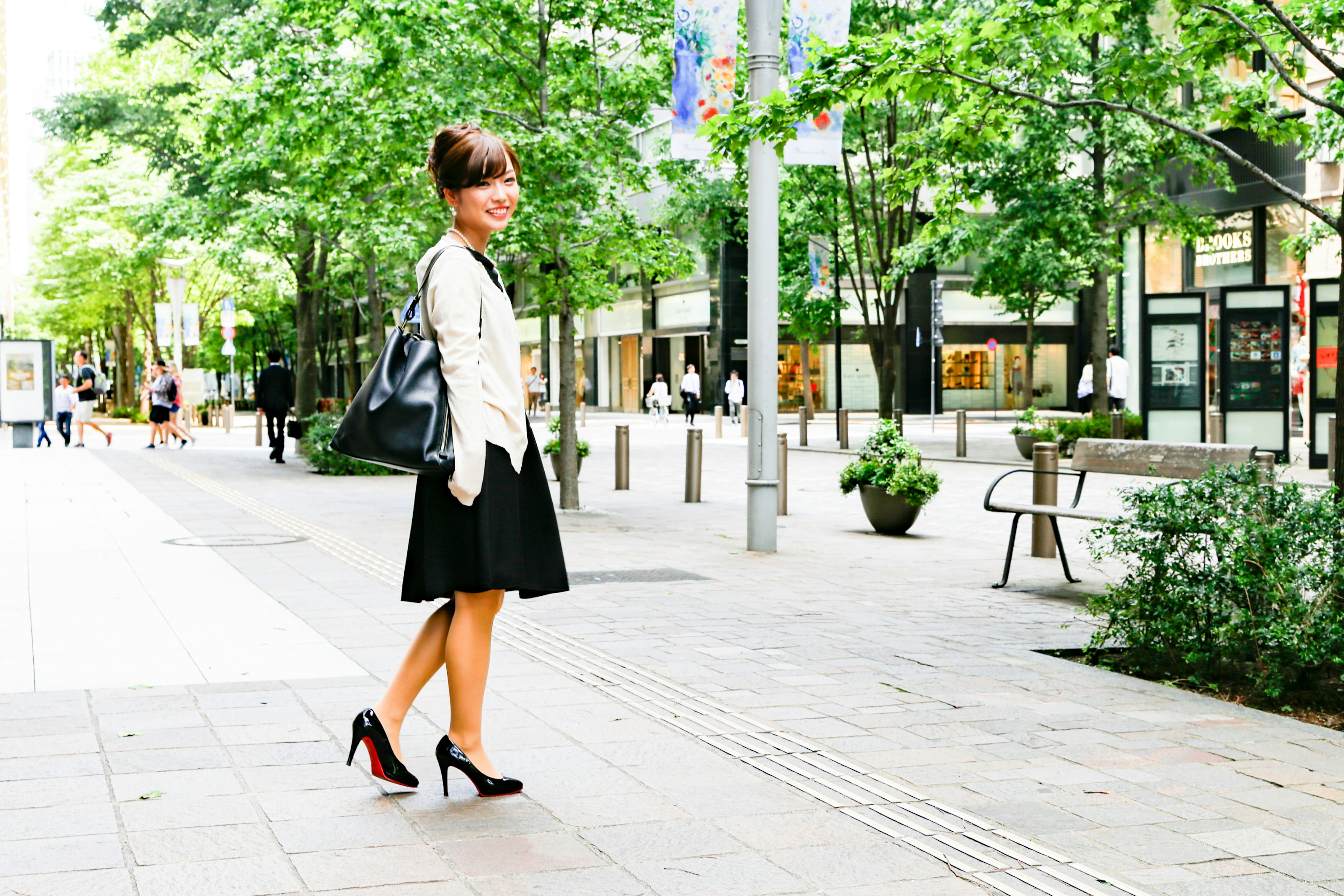 Woman walking in a park wearing a short skirt and high heels holding a bag green trees in the background