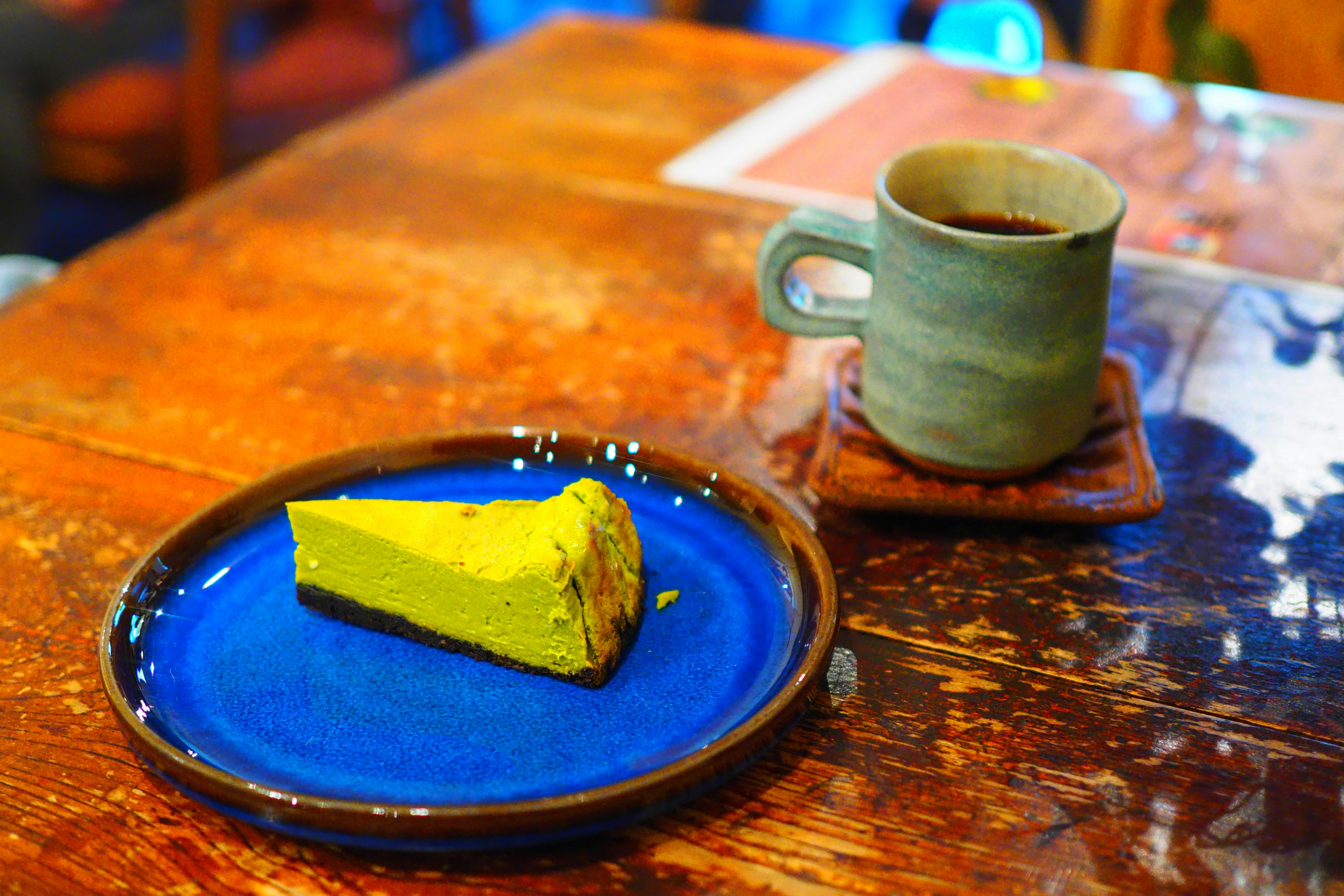 Sepotong kue kuning di piring biru di samping cangkir abu-abu kopi di atas meja kayu