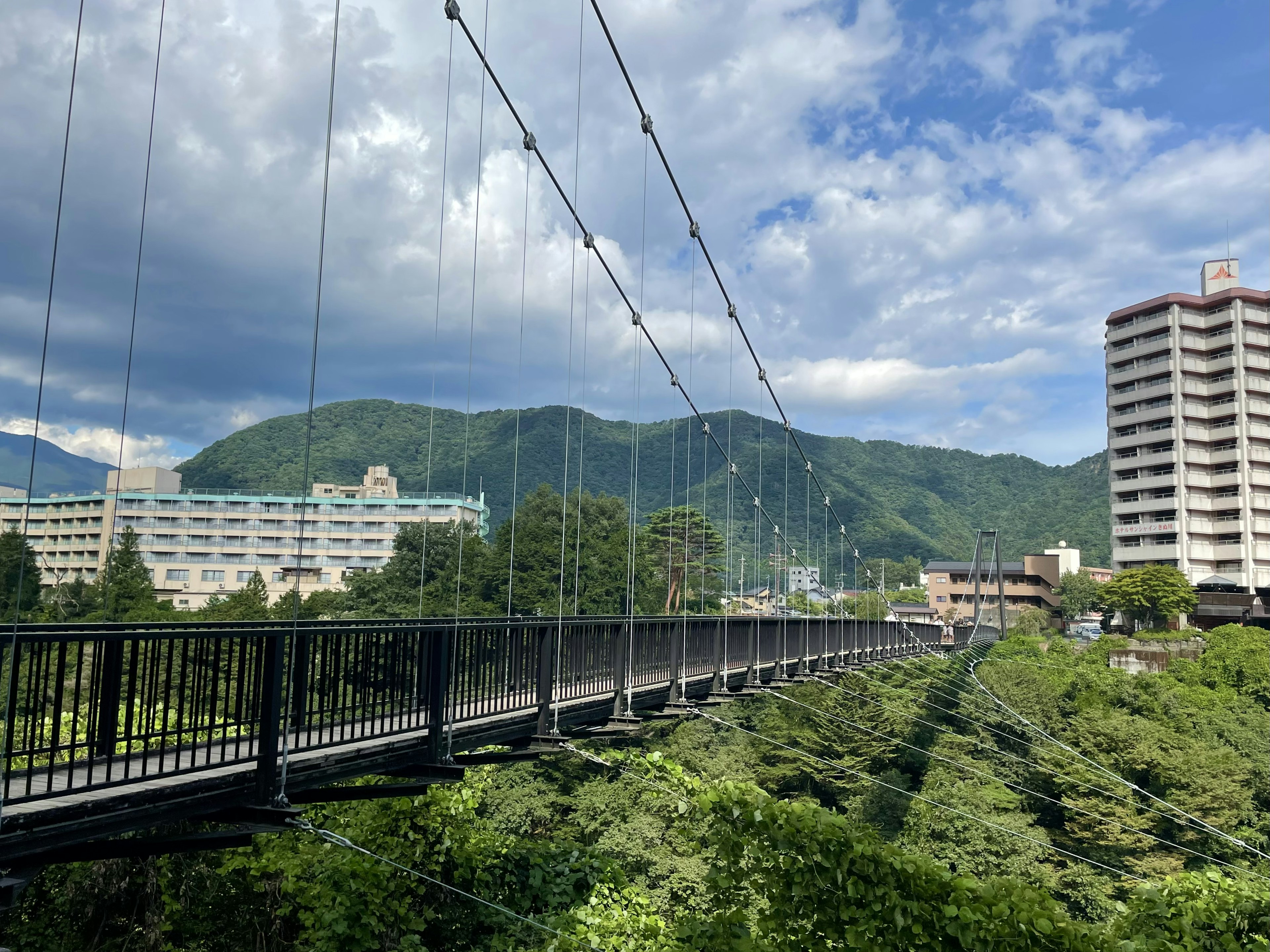 美しい吊り橋が青い空の下にあり、周囲には緑豊かな山々と建物が見える