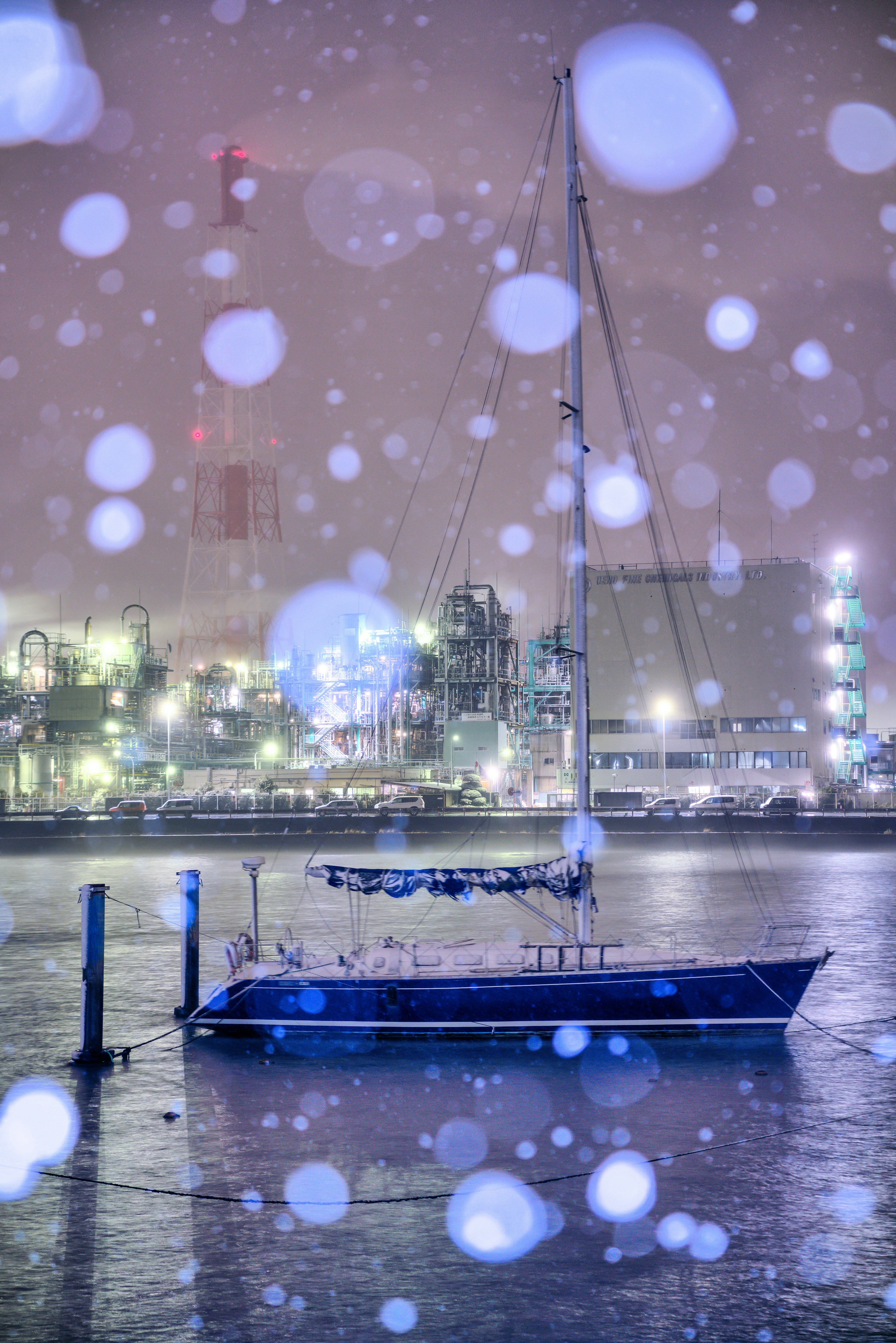 雪の降る夜に港に停泊している青いヨットと背景の工場