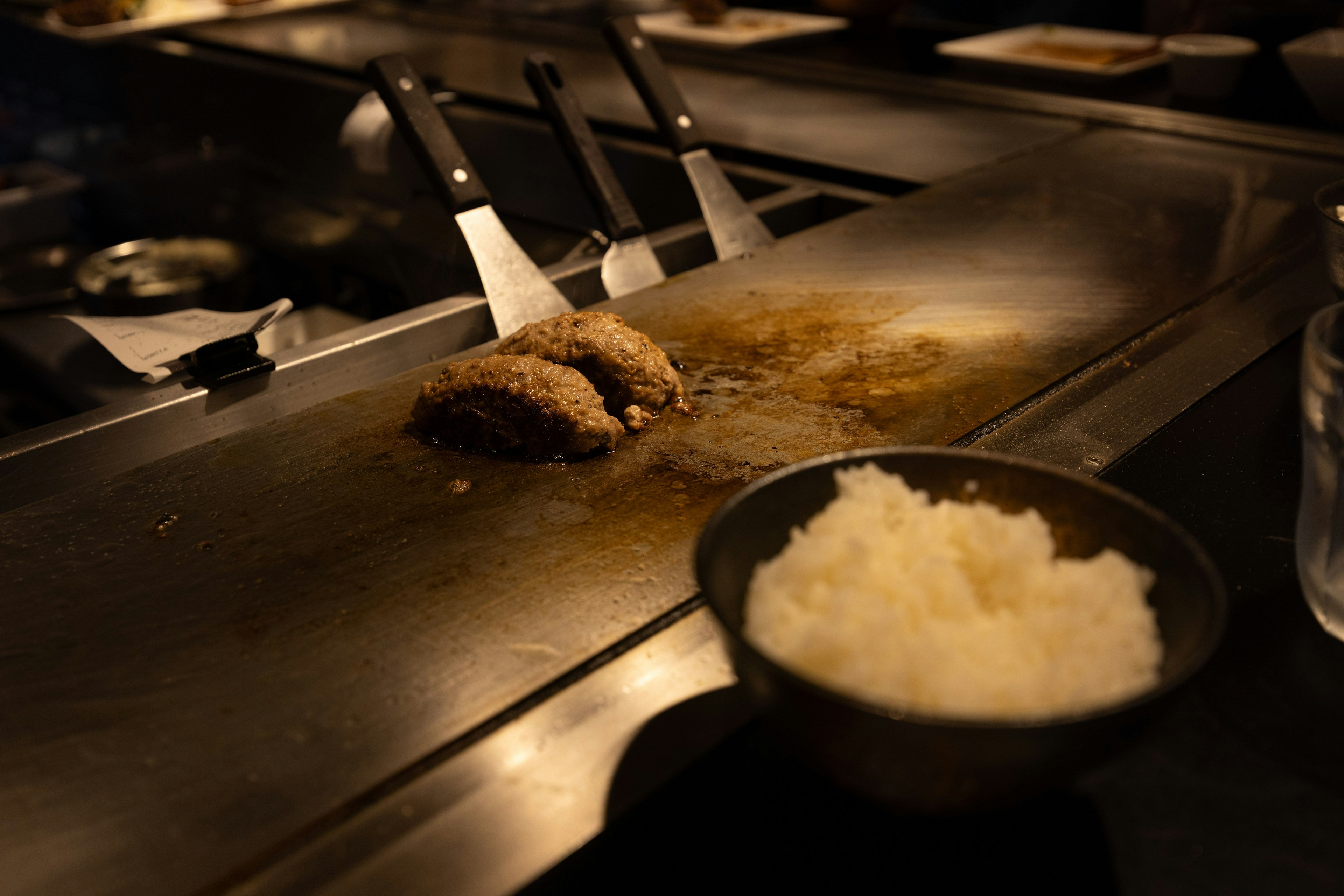 Eine Szene mit gegrillten Hamburger-Patties auf einer Teppan und einer Schüssel weißen Reis