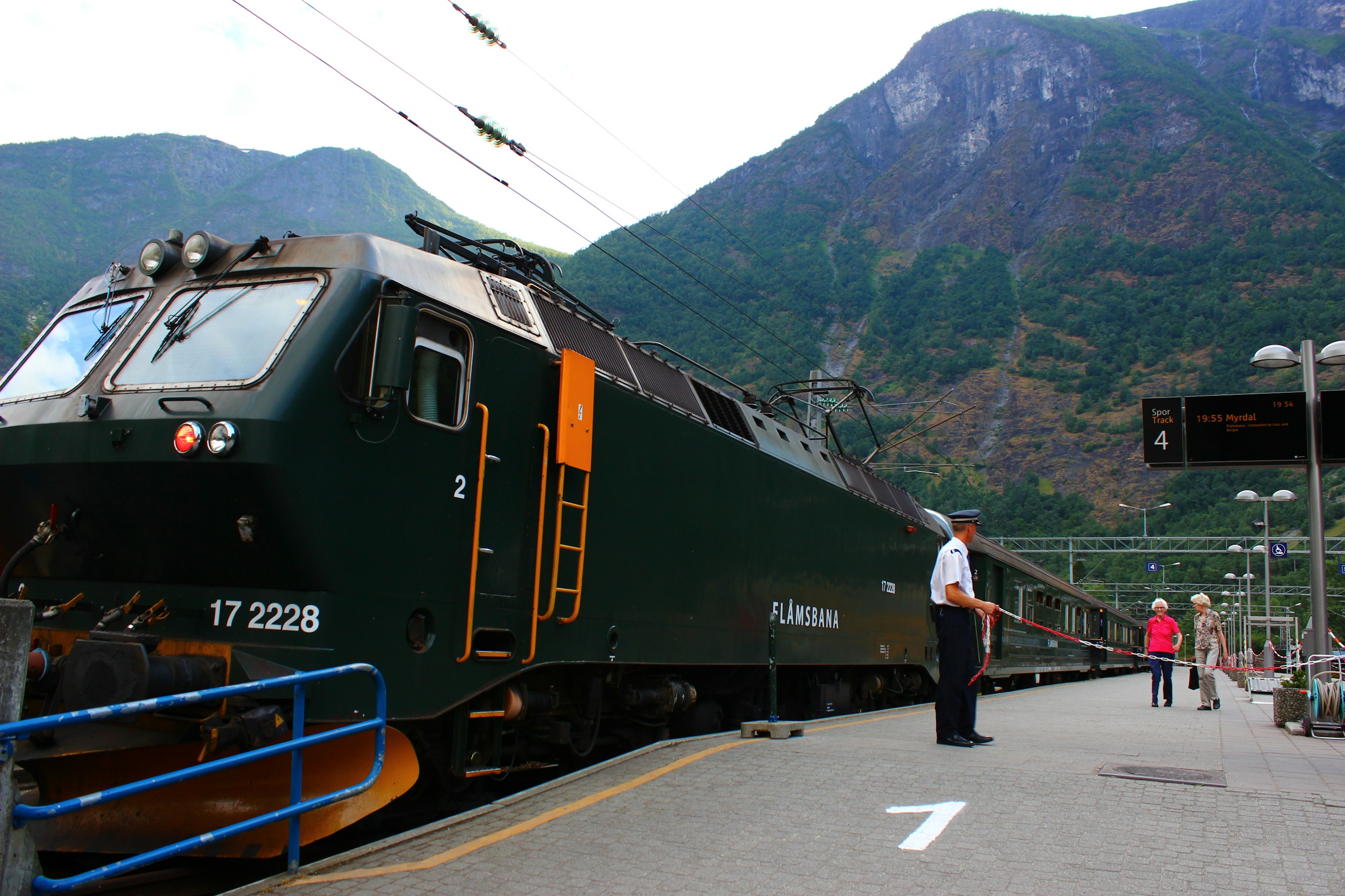 Un treno verde in una stazione con passeggeri che scendono e montagne sullo sfondo