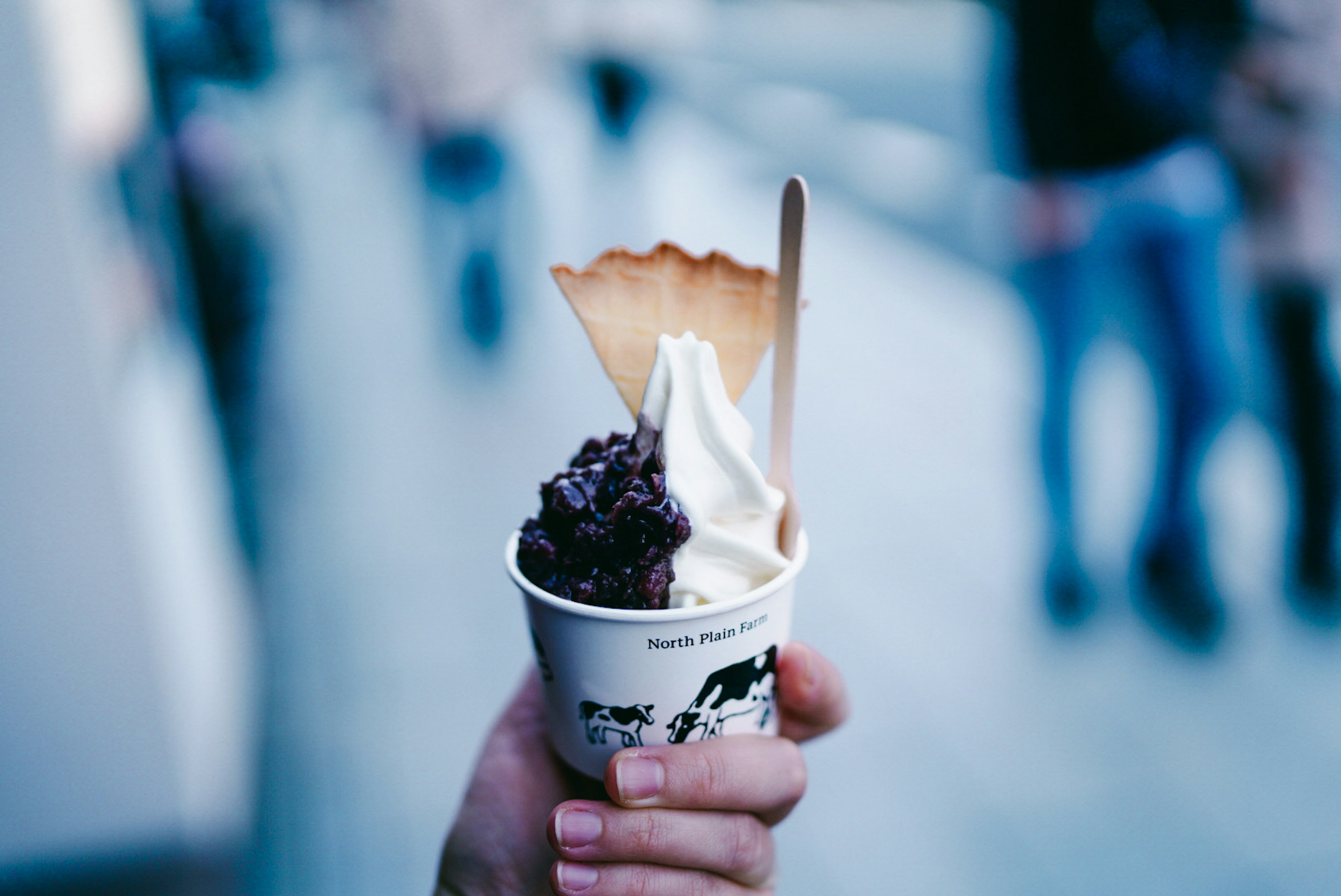 Taza de helado sostenida en la mano con helado oscuro y crema batida coronada con un cono en un entorno callejero borroso