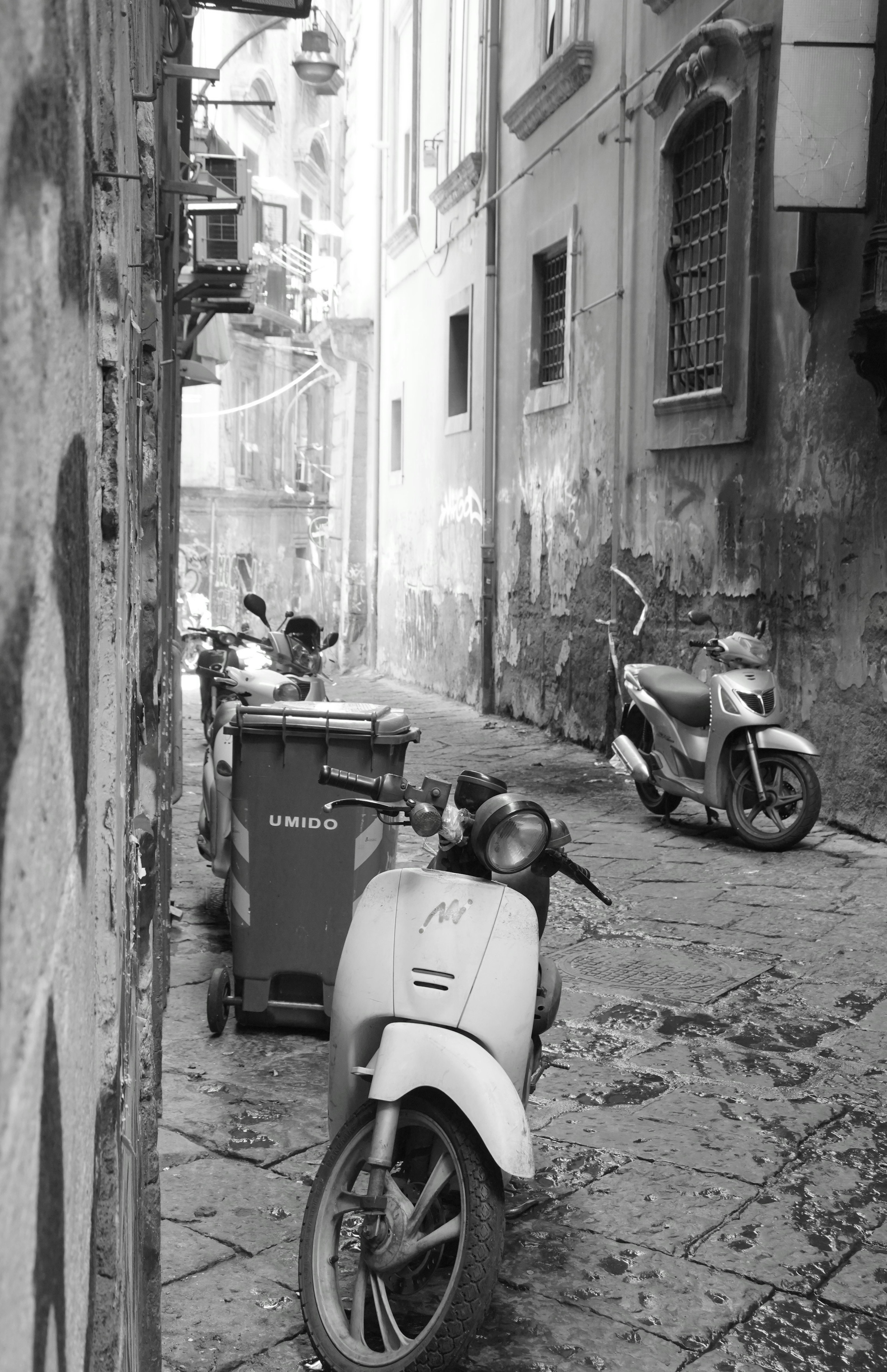 Narrow alley with parked scooters and trash bins in black and white