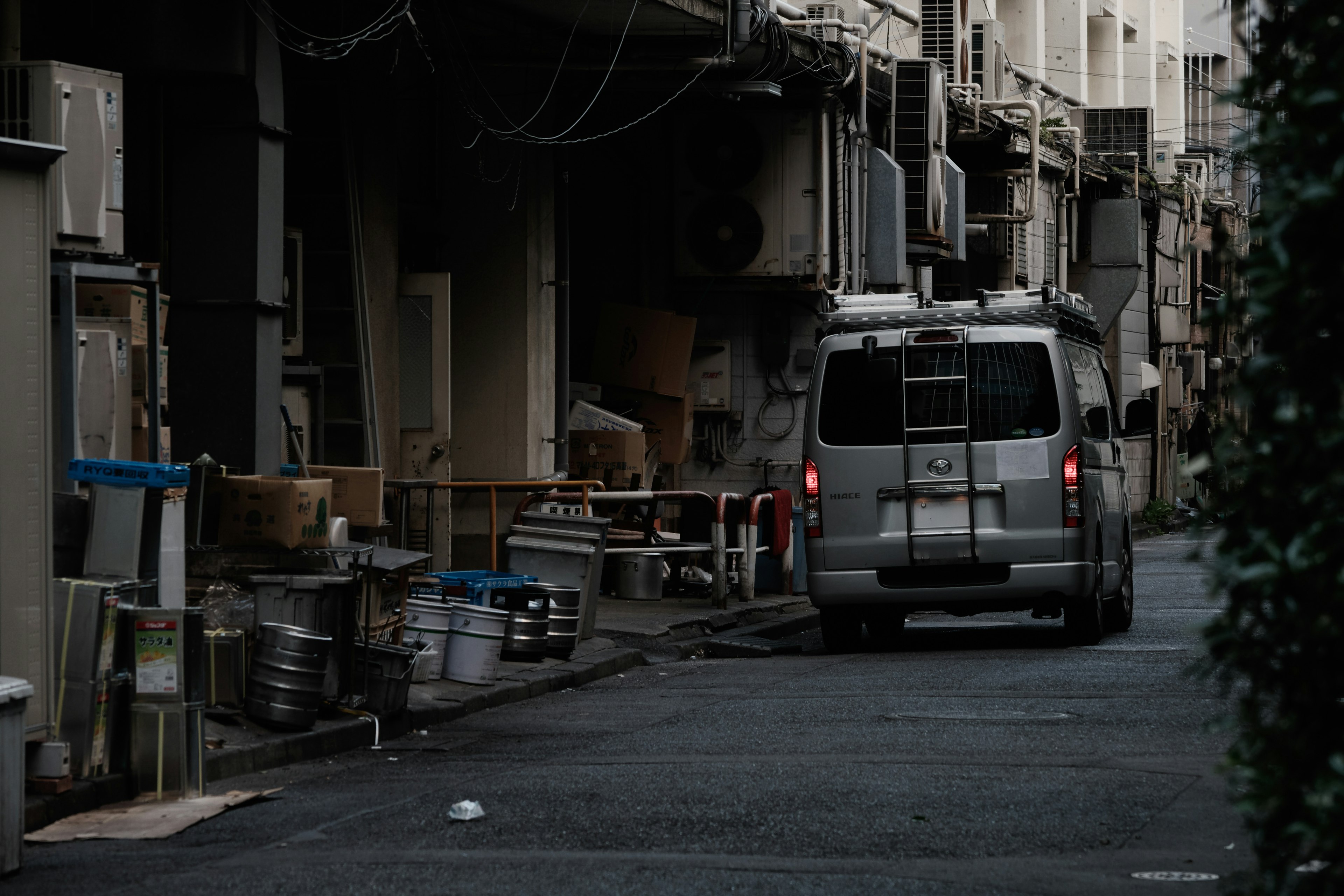 狭い路地に停まっている銀色のバンと周囲の物置