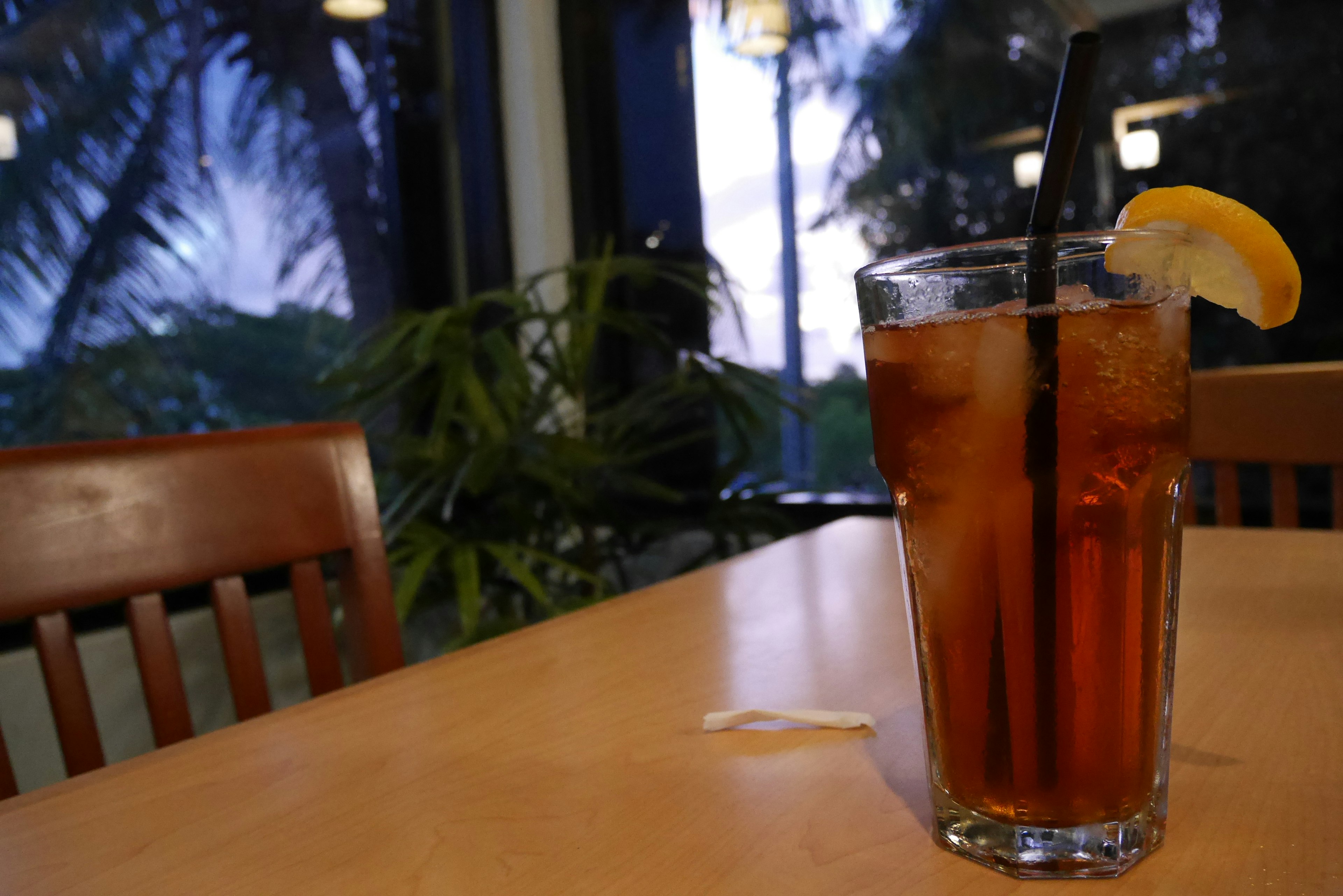 Un vaso de té helado en una mesa con una rodaja de limón y una pajita palmeras visibles fuera de la ventana