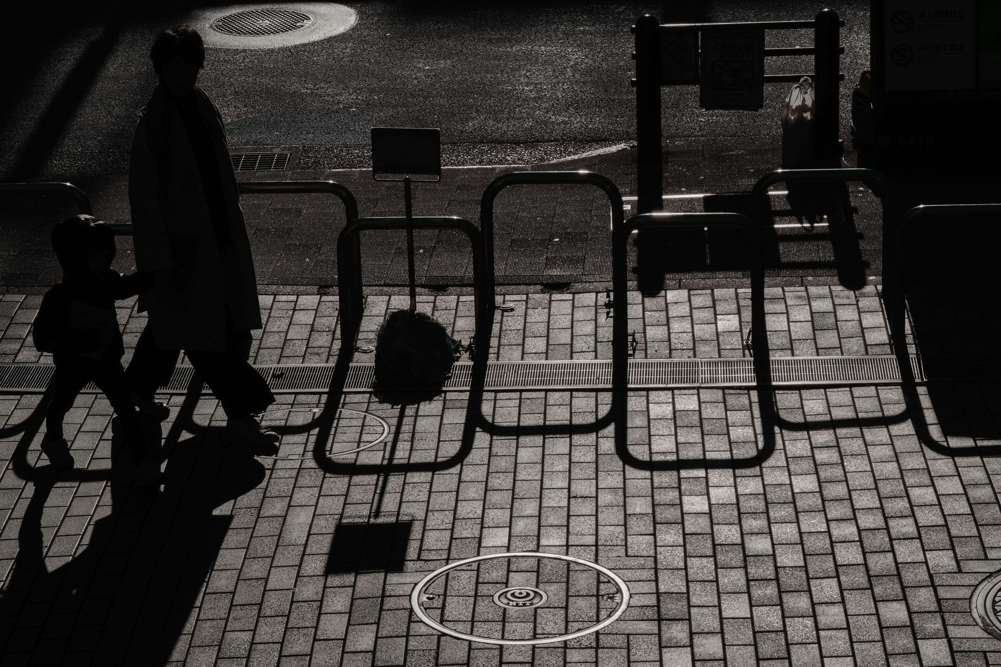 Silhouette of a person walking with a child near barriers in a black and white urban scene