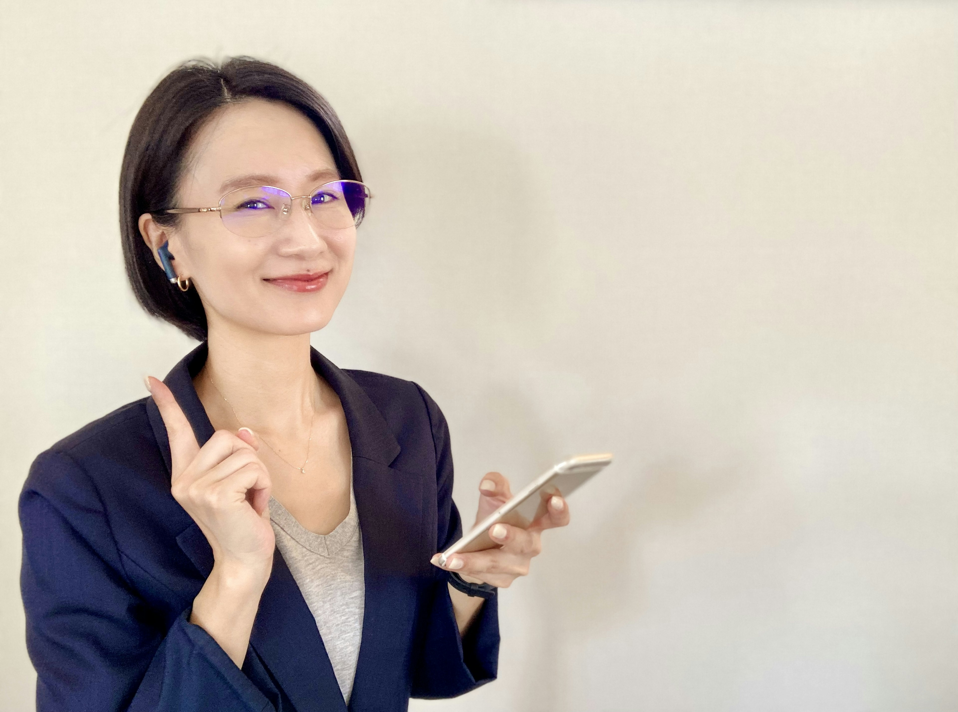 Mujer de negocios sonriendo y sosteniendo un teléfono inteligente con un dedo levantado frente a una pared blanca