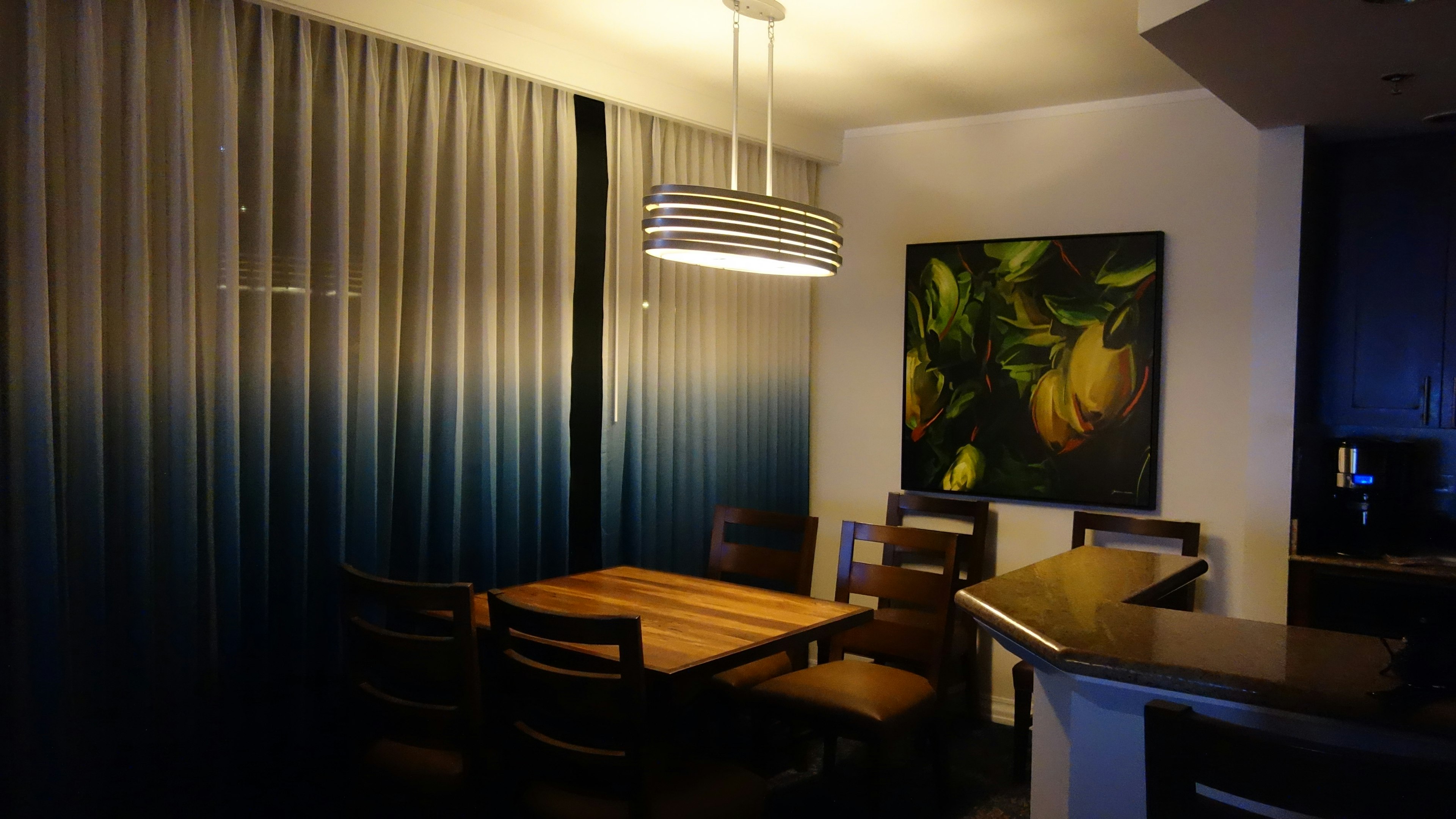 Interior de comedor con cortinas de degradado azul a blanco mesa y sillas de madera iluminación moderna y una pintura de frutas en la pared