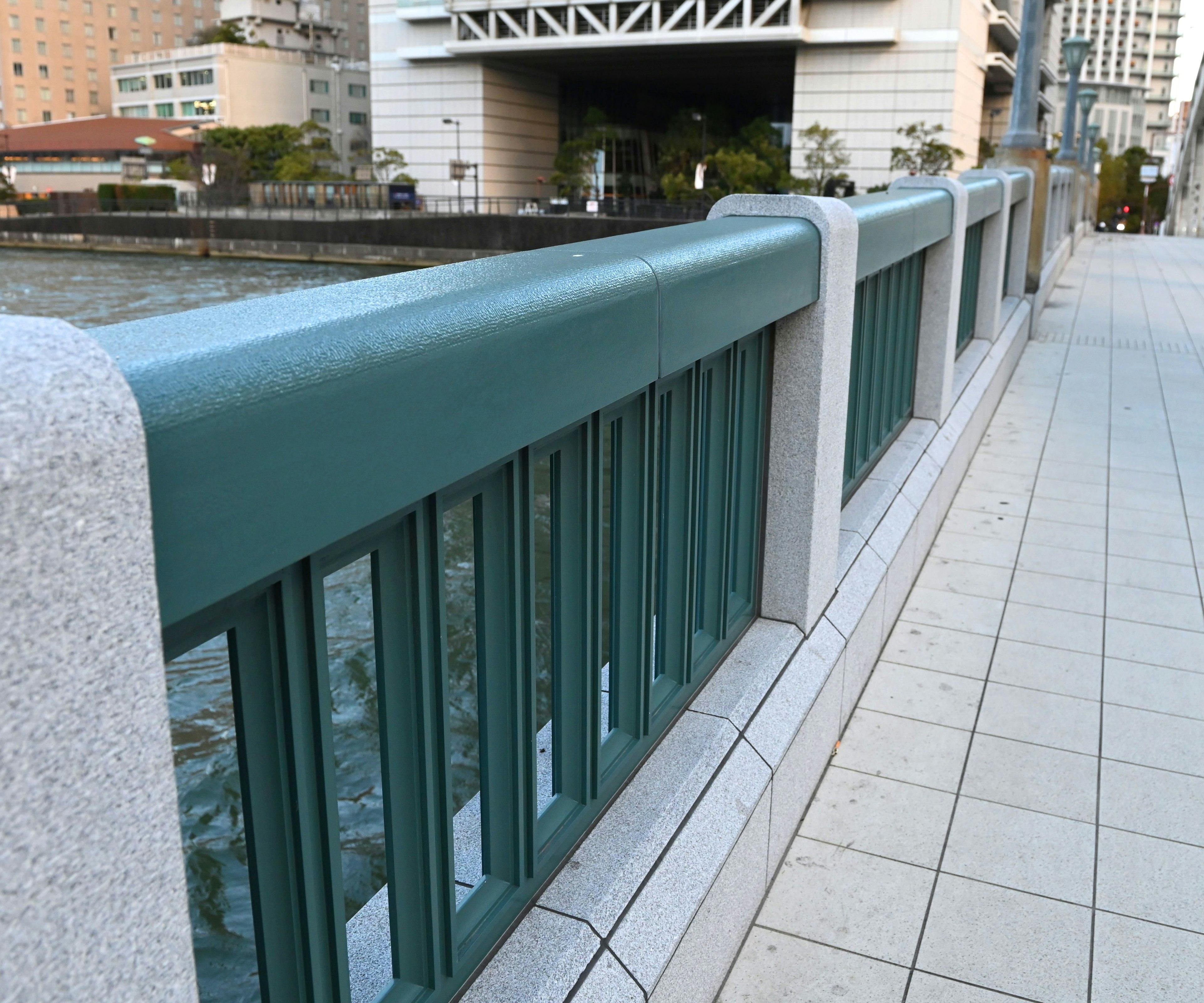 Foto de una barandilla de puente en verde azulado con una acera de baldosas al lado