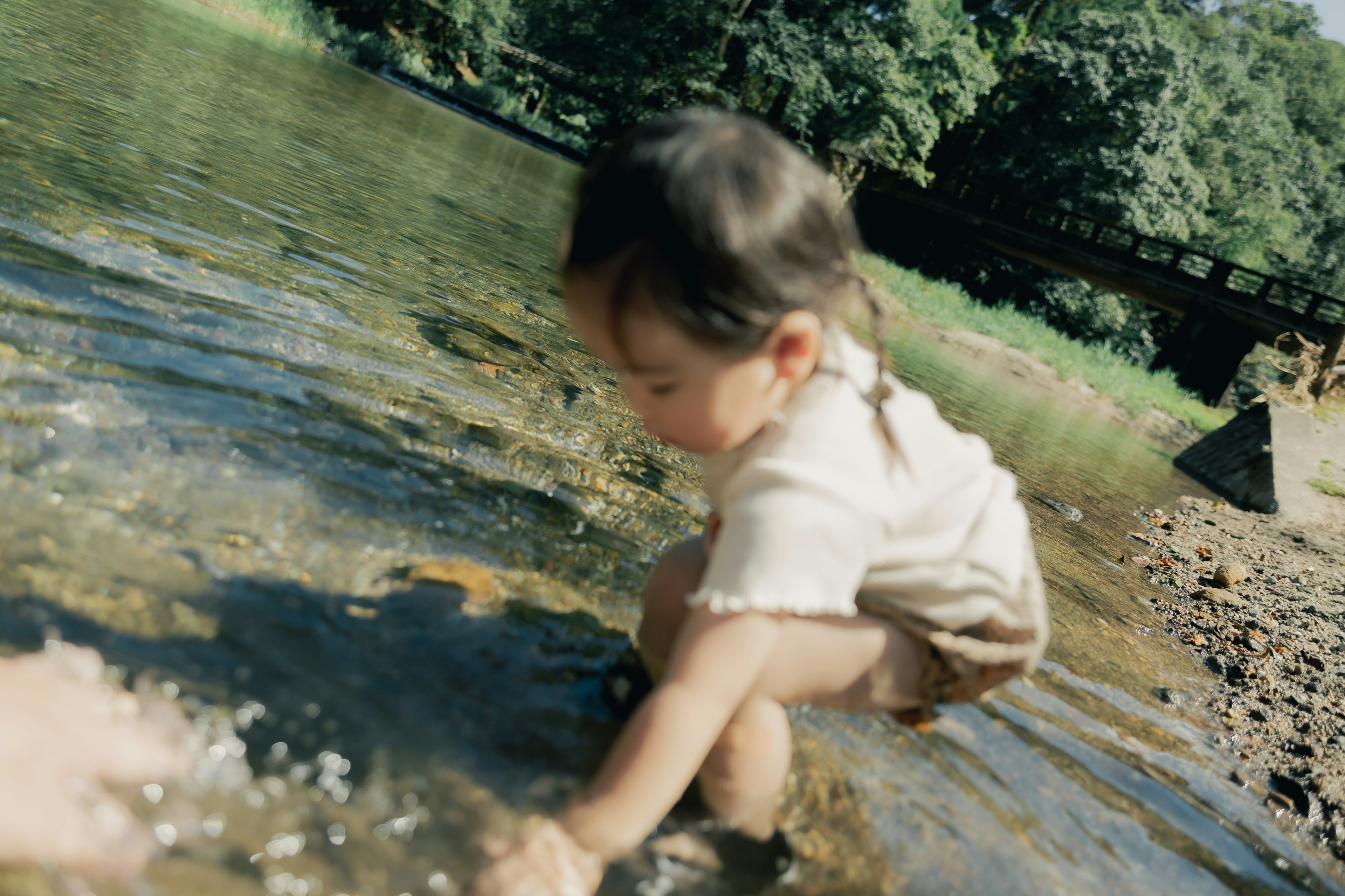 Ein kleines Kind spielt am Wasser mit grünen Bäumen und einer Brücke im Hintergrund