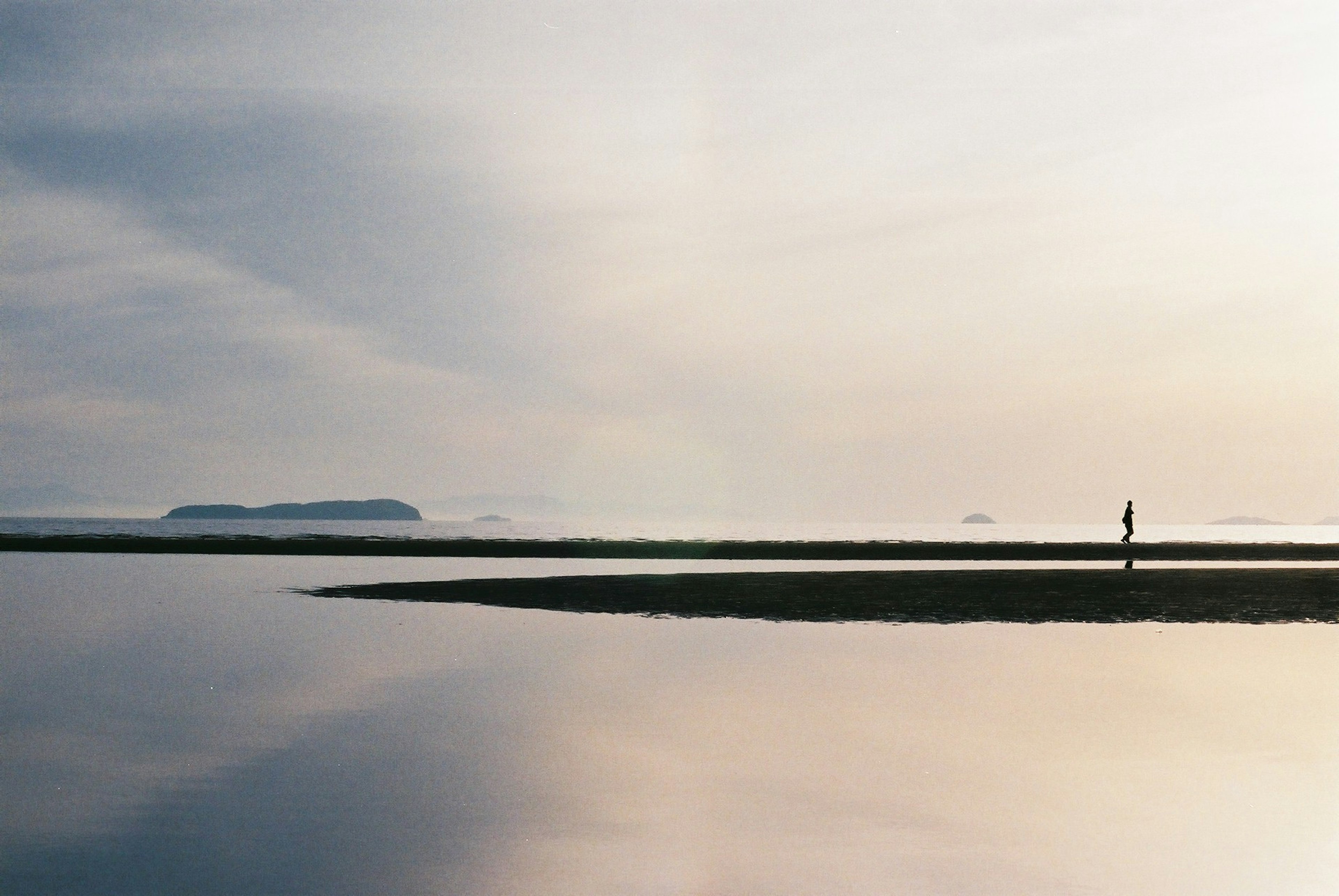 Silhouette einer Person, die an einem ruhigen Strand mit ruhigem Wasser geht, das den Himmel reflektiert