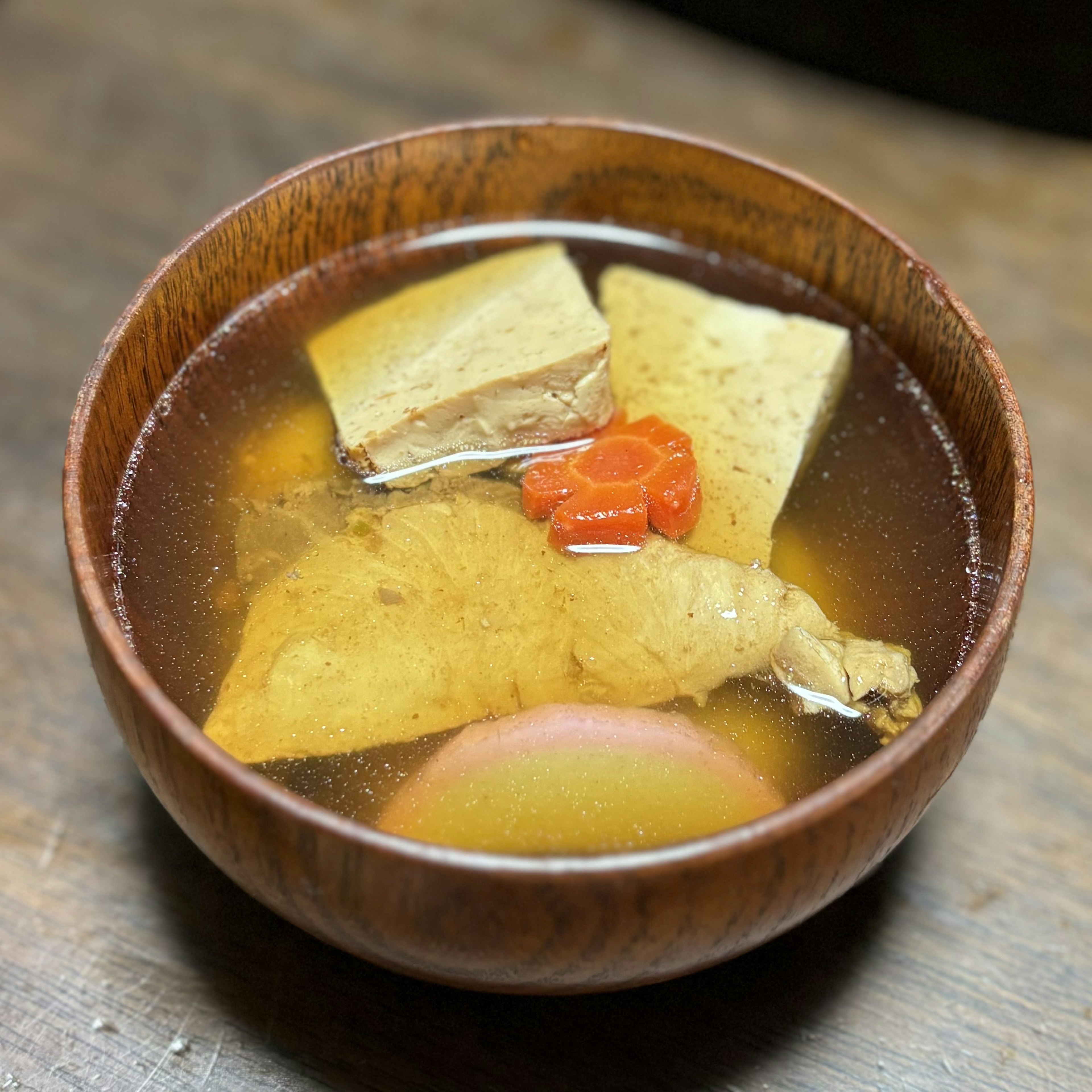 Una ciotola di legno piena di zuppa con pollo, tofu e ingredienti erboristici