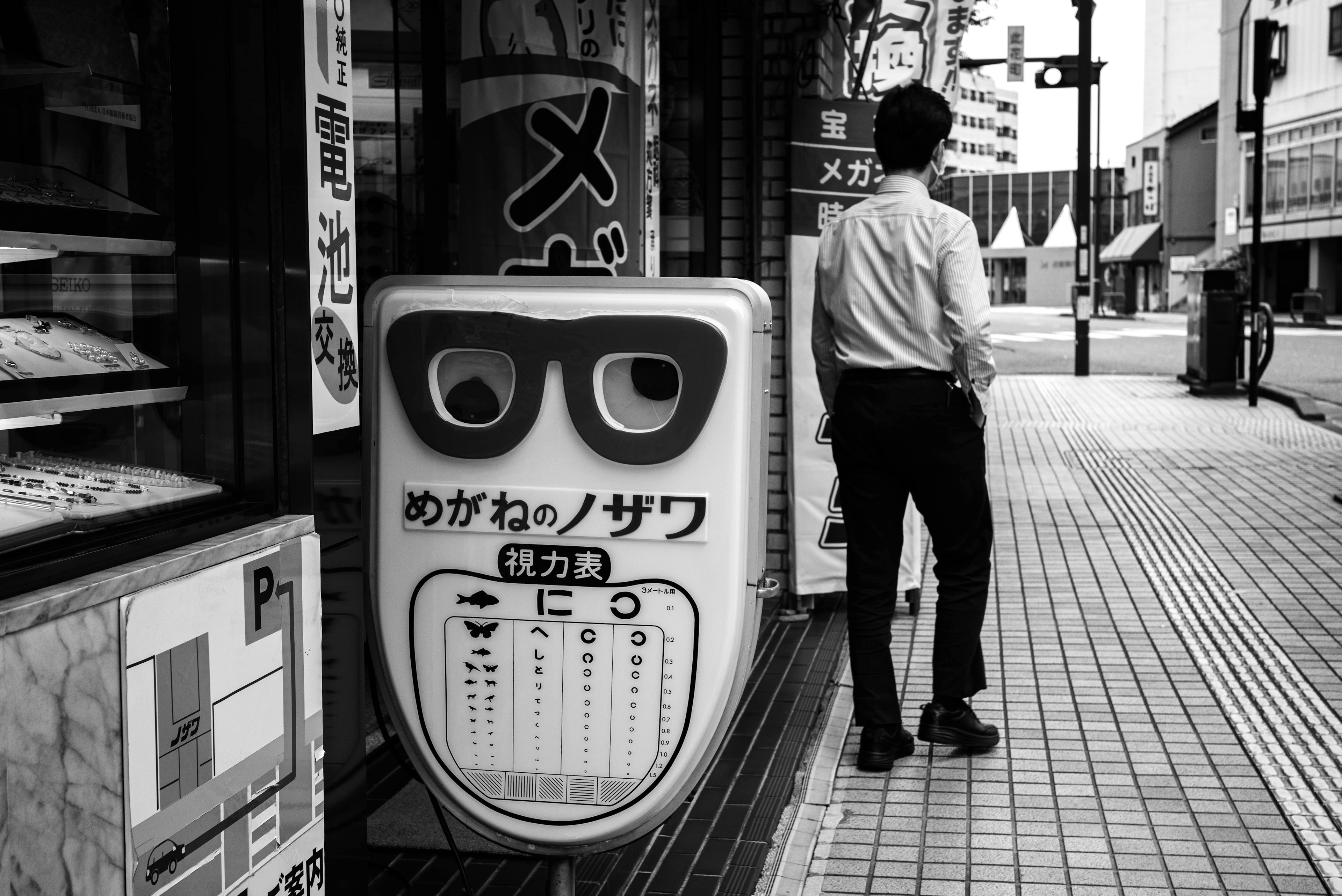 白黒の街並みに立つ男性と眼鏡の形をした看板が特徴的な風景