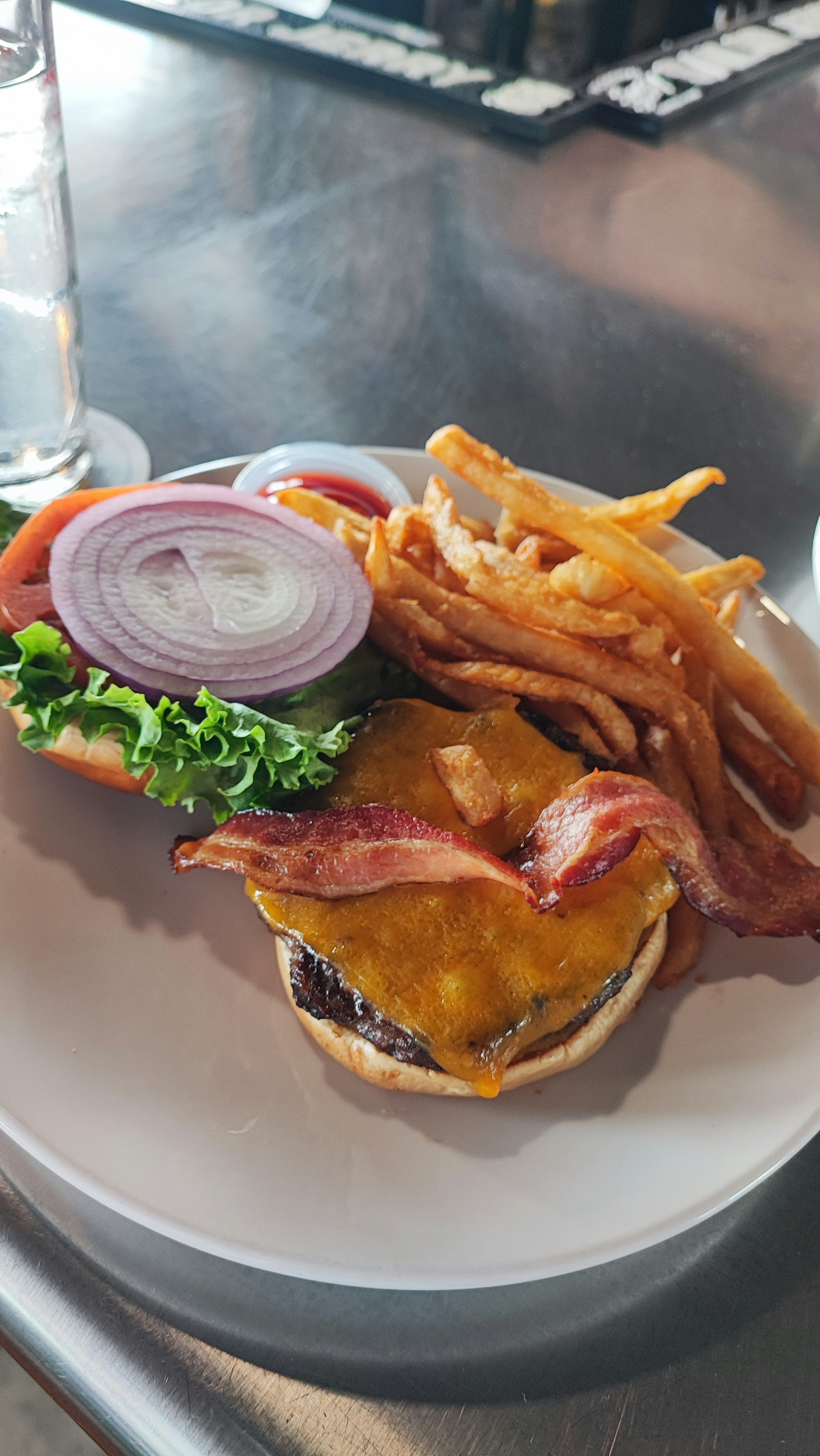 Un plato con una hamburguesa con queso cubierta de tocino lechuga tomate y rodajas de cebolla roja acompañado de papas fritas en espiral