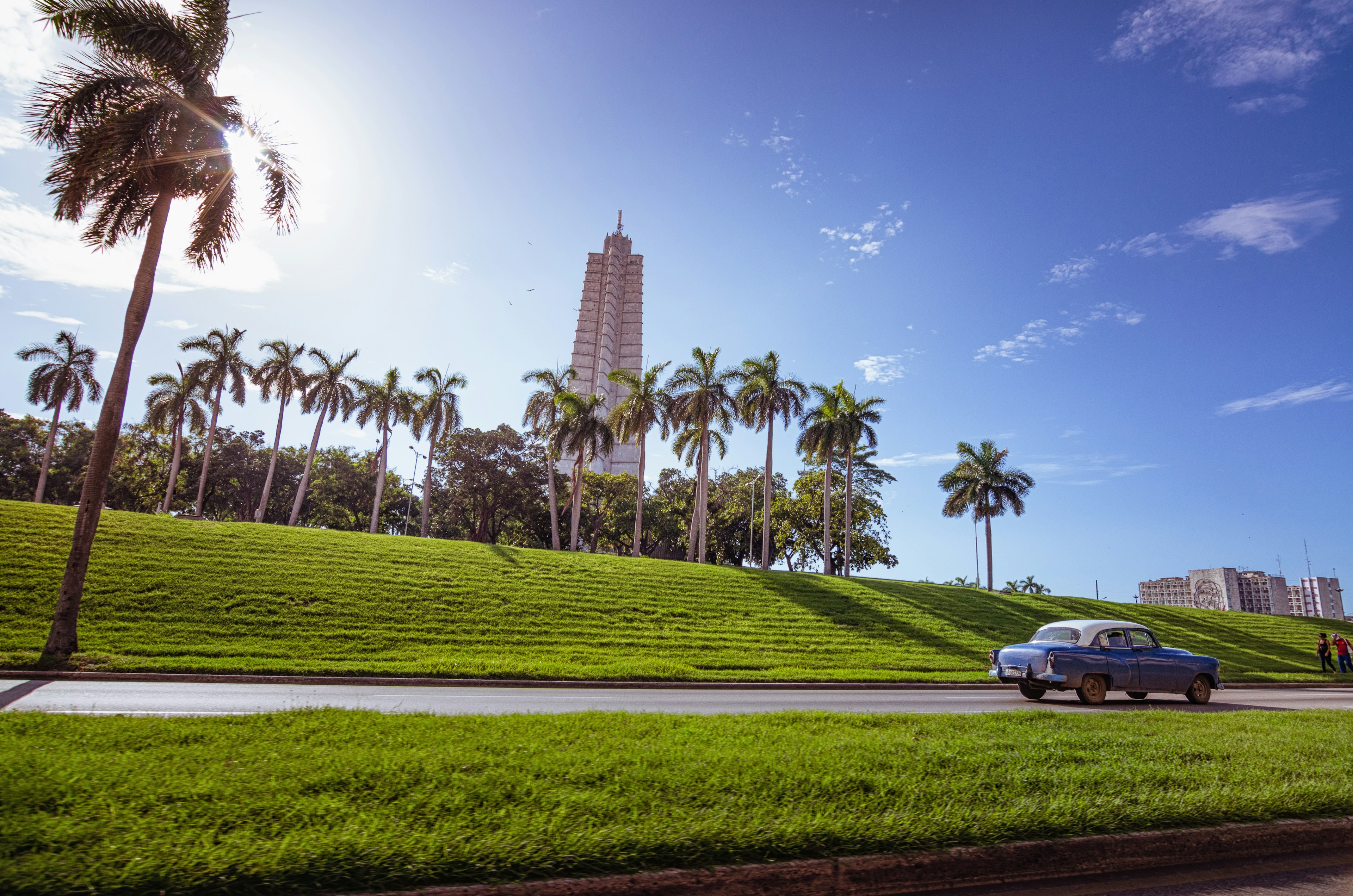 Scenic view featuring a tall building and palm trees under a blue sky with a vintage car on the road