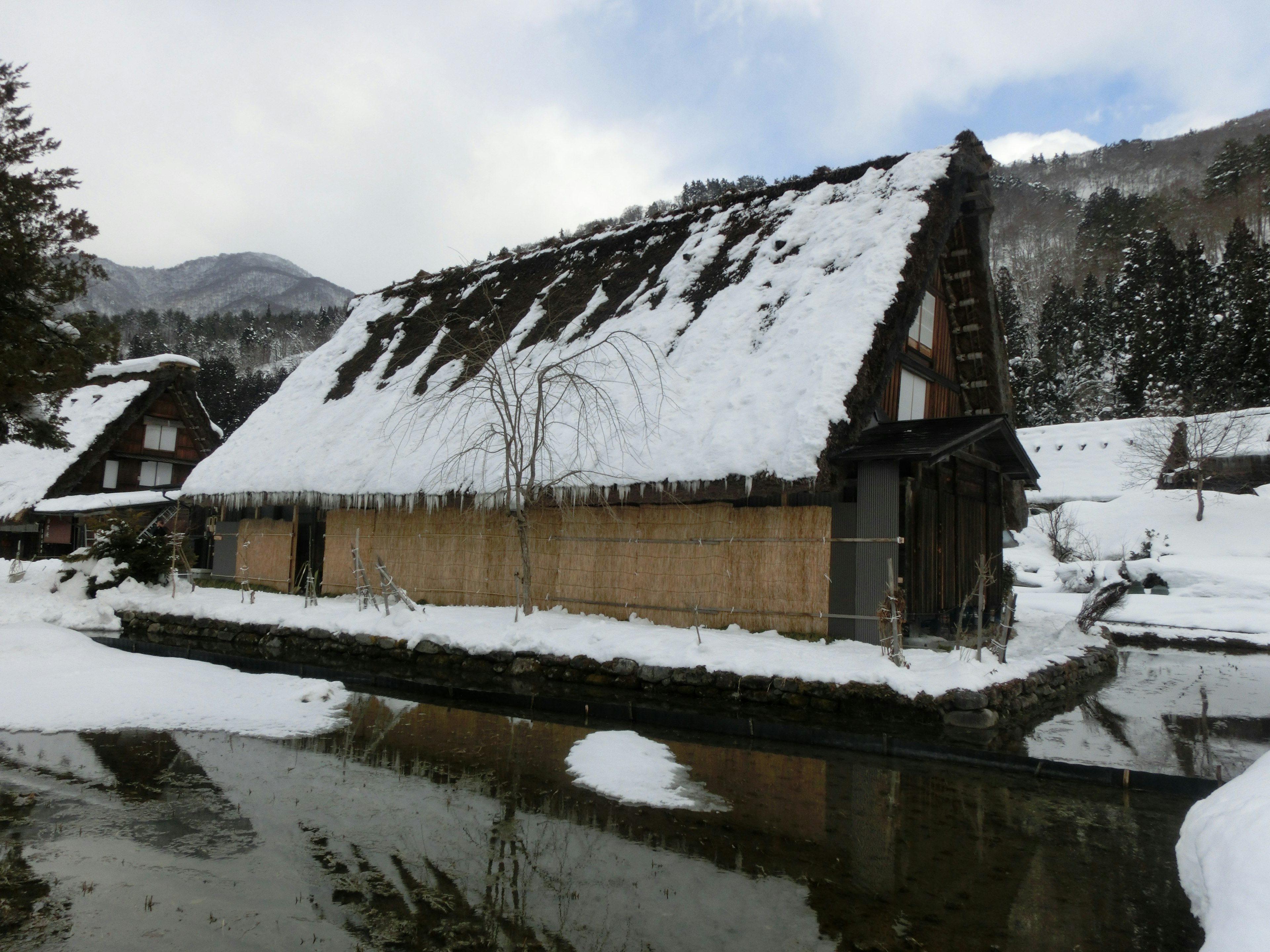 Snow-covered gassho-zukuri house next to a calm water surface