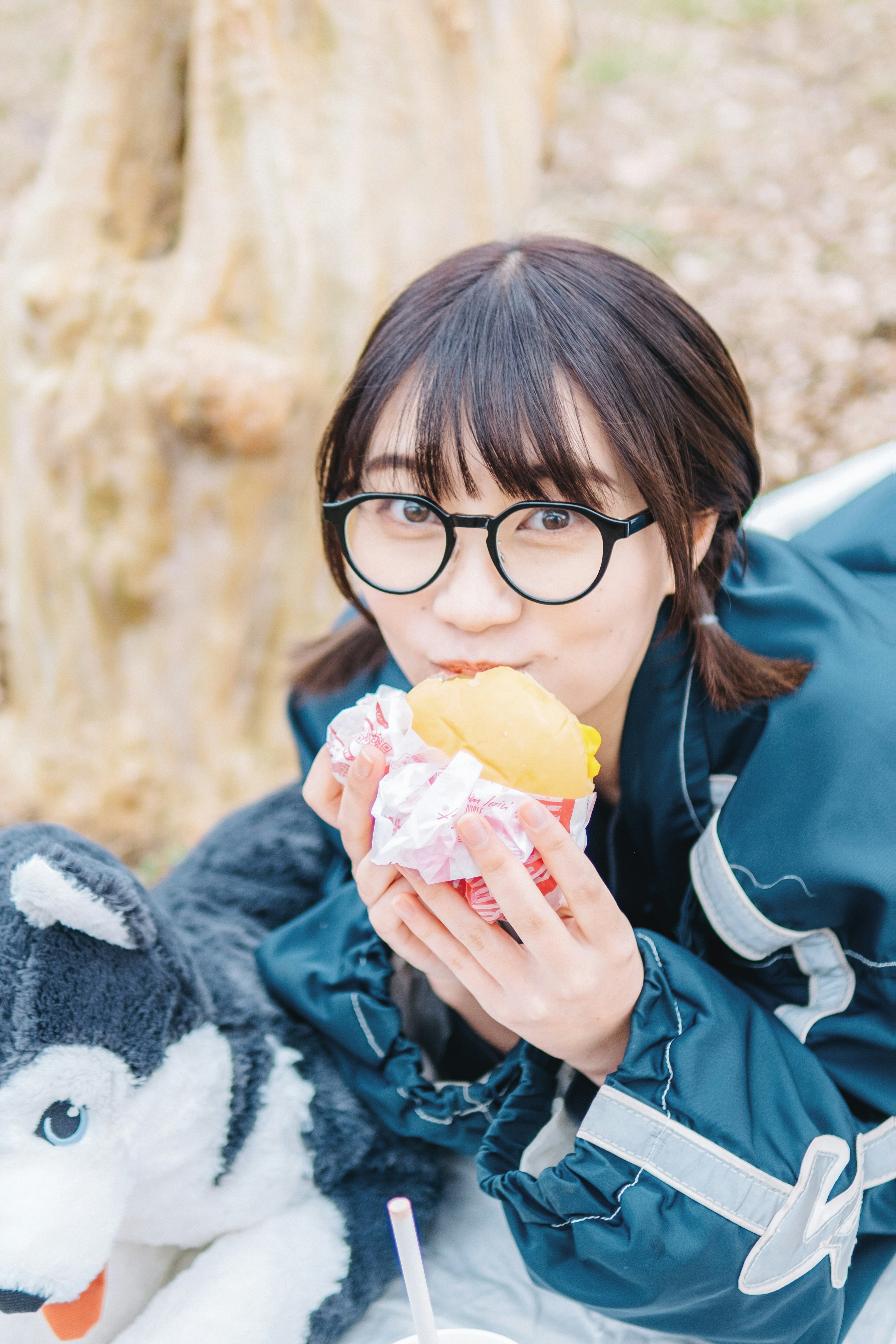 Chica comiendo un sándwich con una chaqueta con capucha gris y un perro husky de peluche cerca