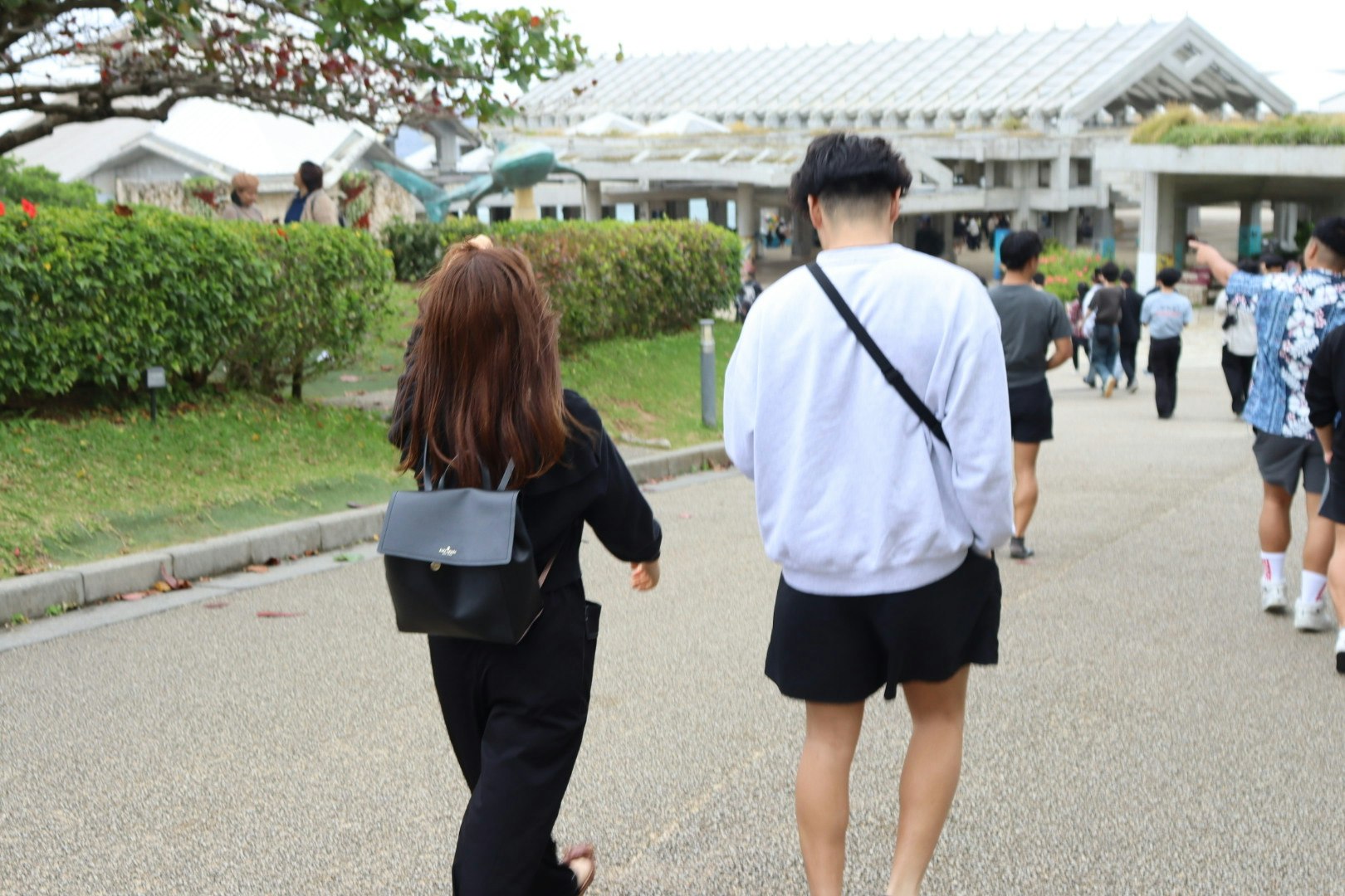 Eine Szene von zwei Personen, die von hinten gehen Eine trägt schwarze Kleidung mit einem kleinen Rucksack und die andere einen weißen Pullover und Shorts