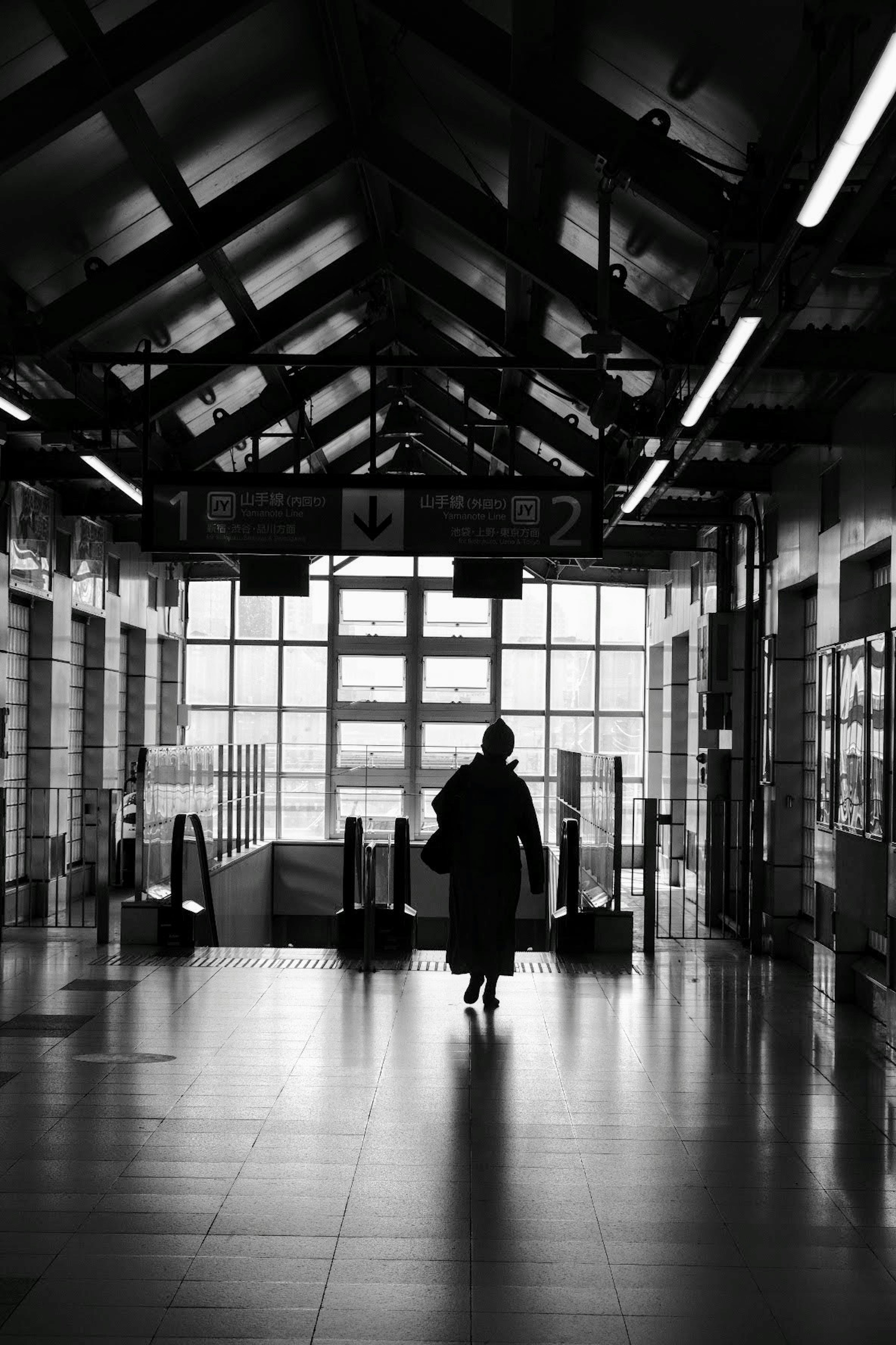 Silhouette seseorang yang berjalan di pintu masuk stasiun dalam Ruangan Cahaya alami yang masuk melalui jendela dalam foto hitam putih