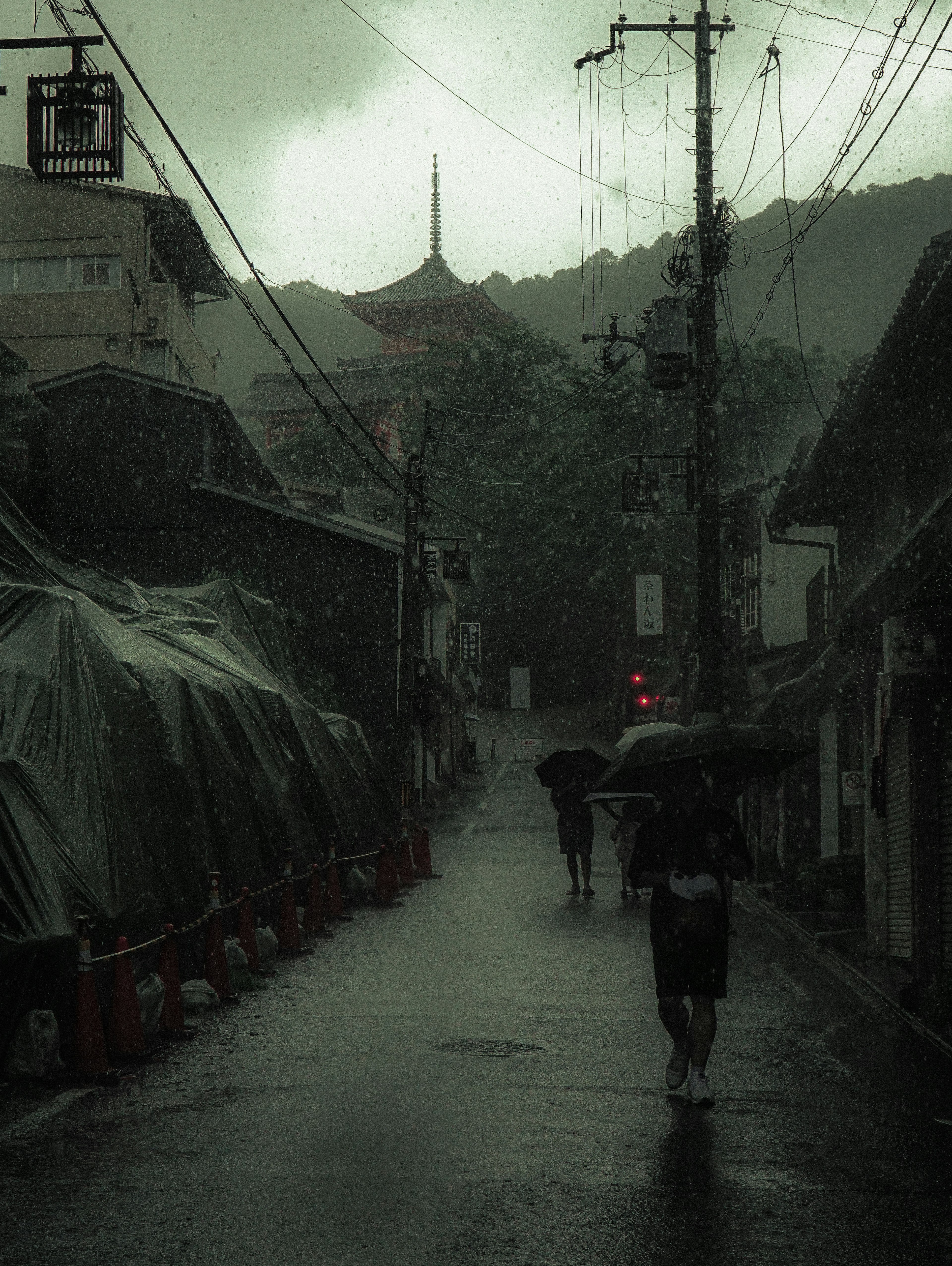 Escena oscura de la calle con personas caminando bajo la lluvia y una torre de templo borrosa al fondo