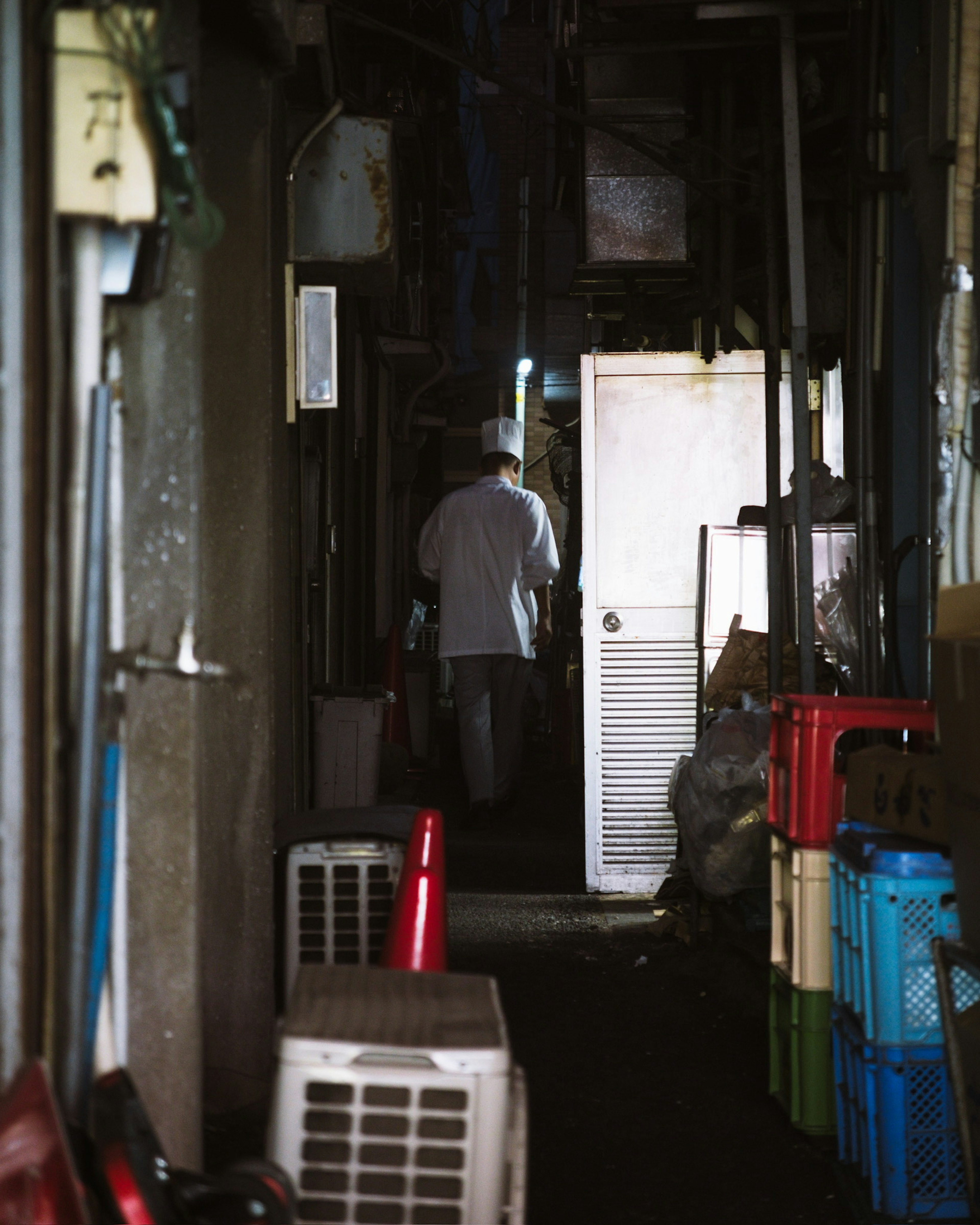 A narrow corridor with a figure facing away and a door in a dimly lit setting