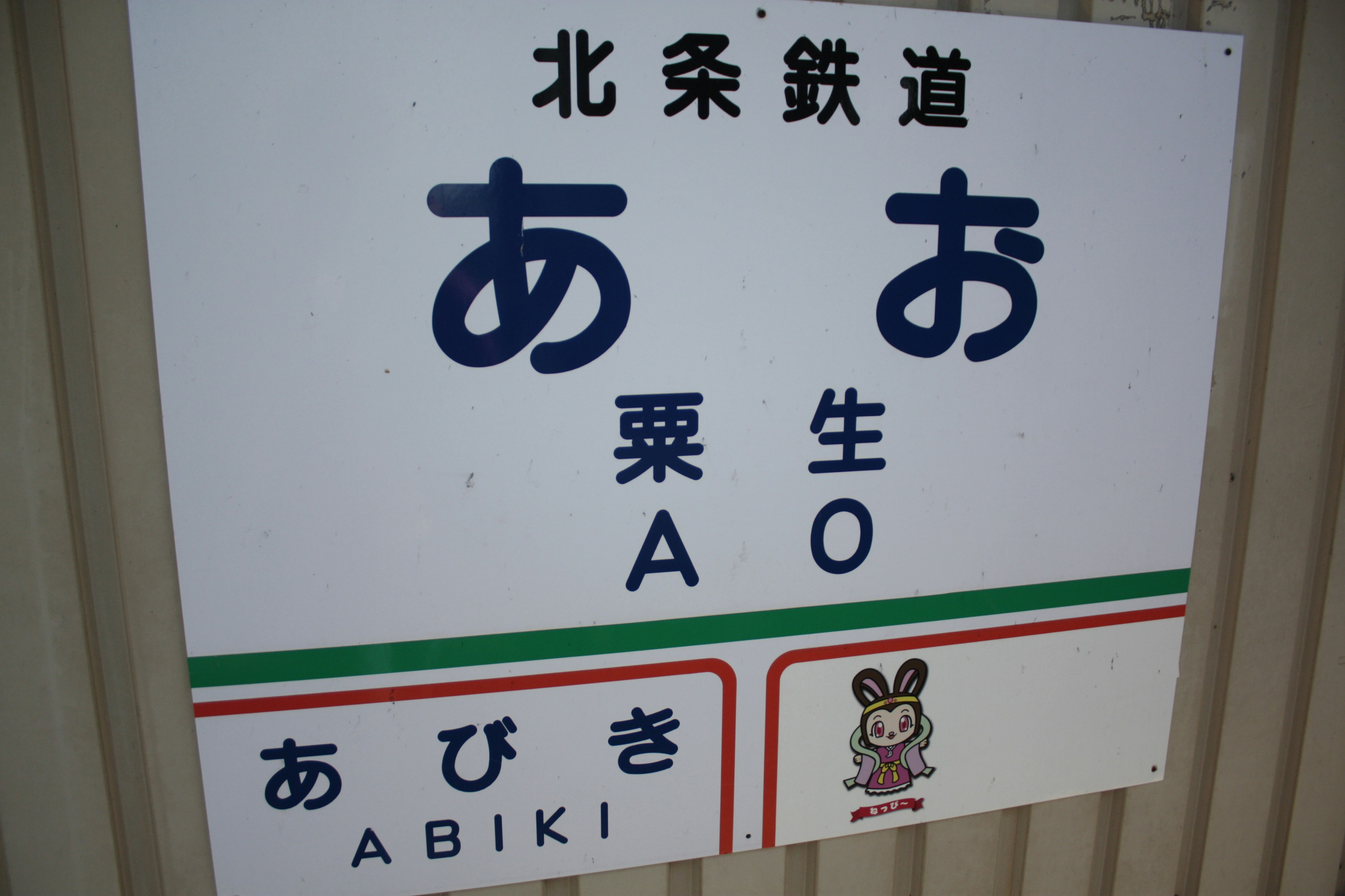 Cartel de la estación Abiki en el ferrocarril Hokuso con los caracteres あお y あびき y una ilustración de dibujos animados