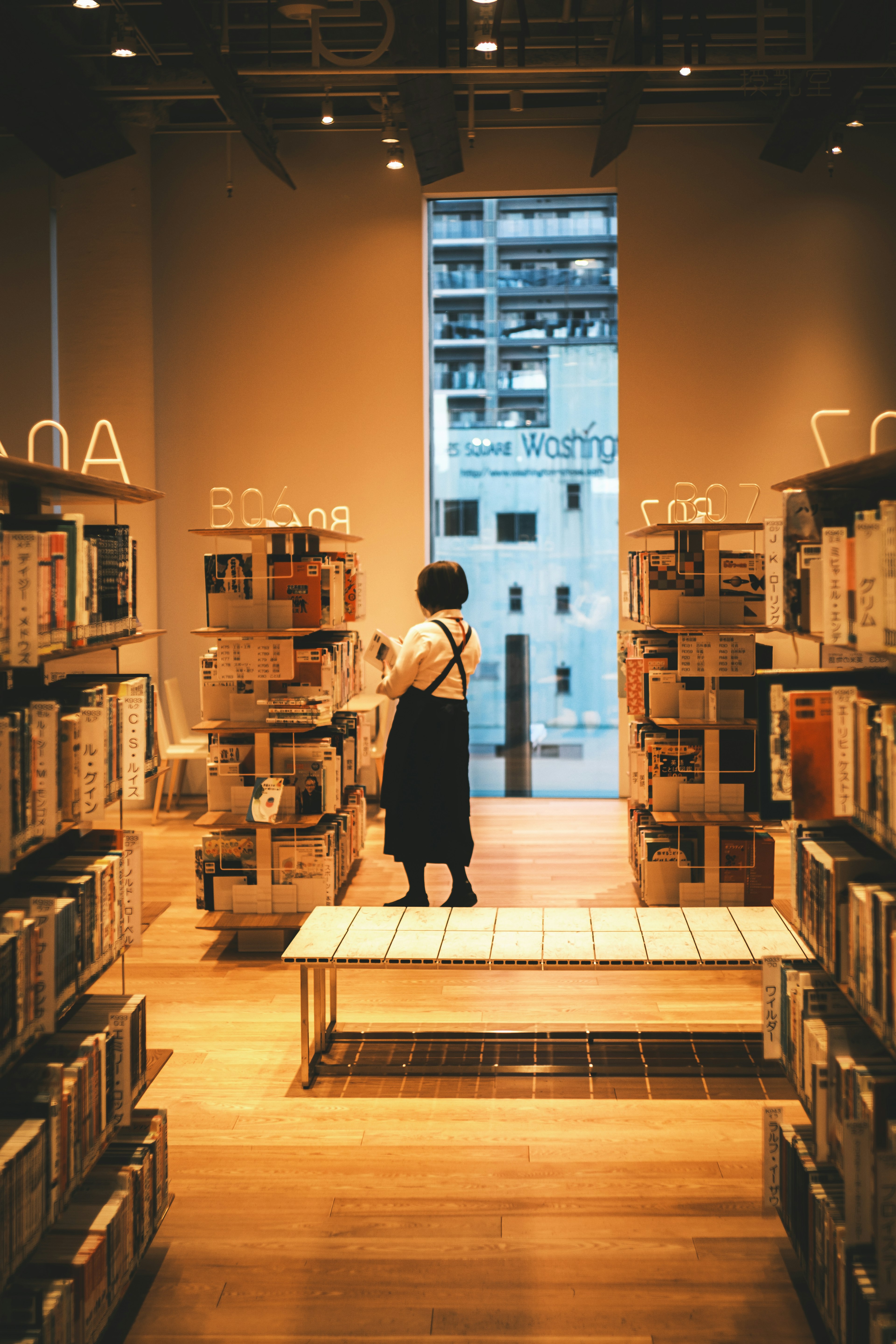 Intérieur d'une librairie avec une femme debout entre des étagères de livres et une vue sur des bâtiments à l'extérieur de la fenêtre