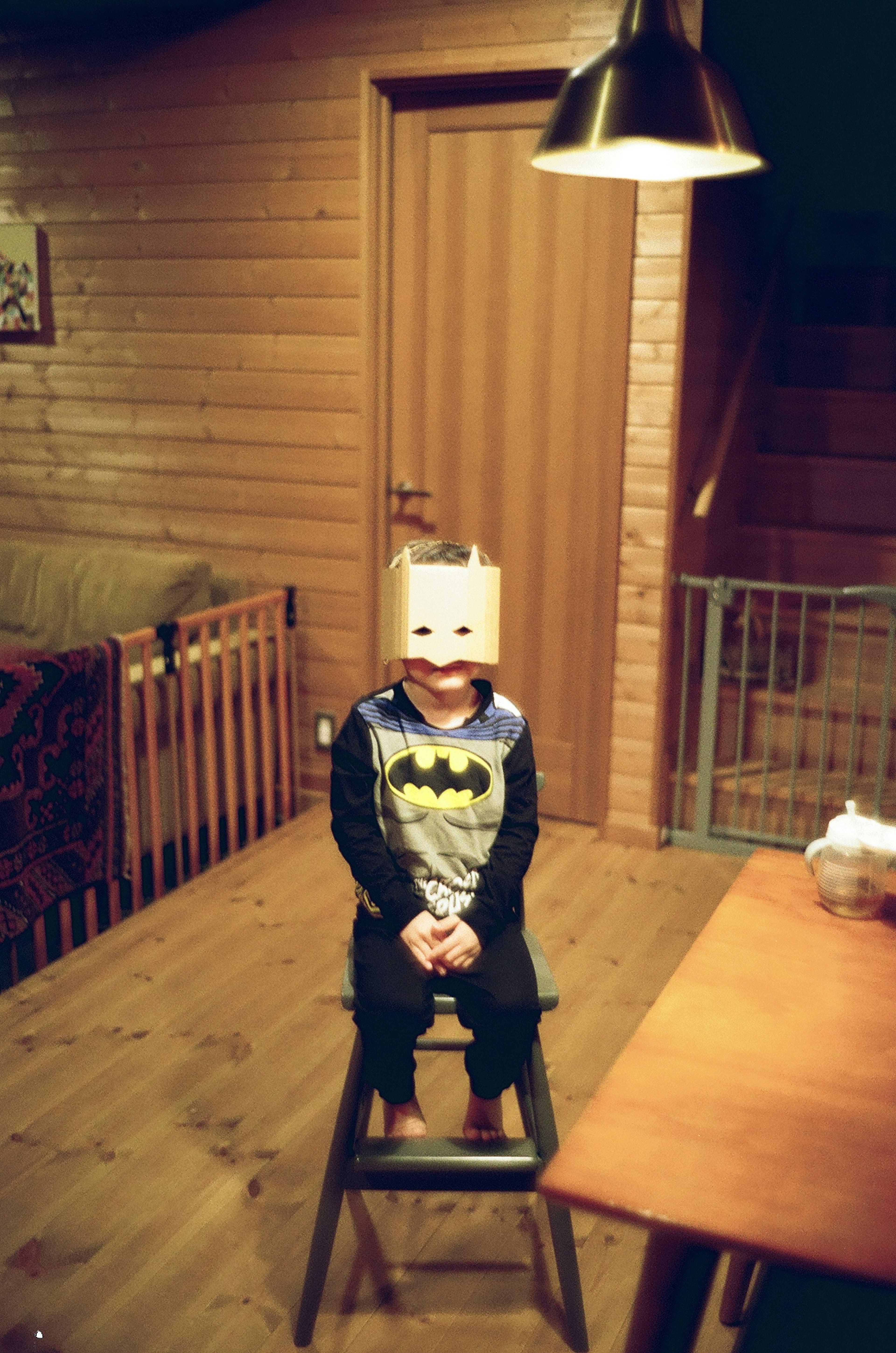 A child wearing a Batman costume sitting on a stool with a box on their head