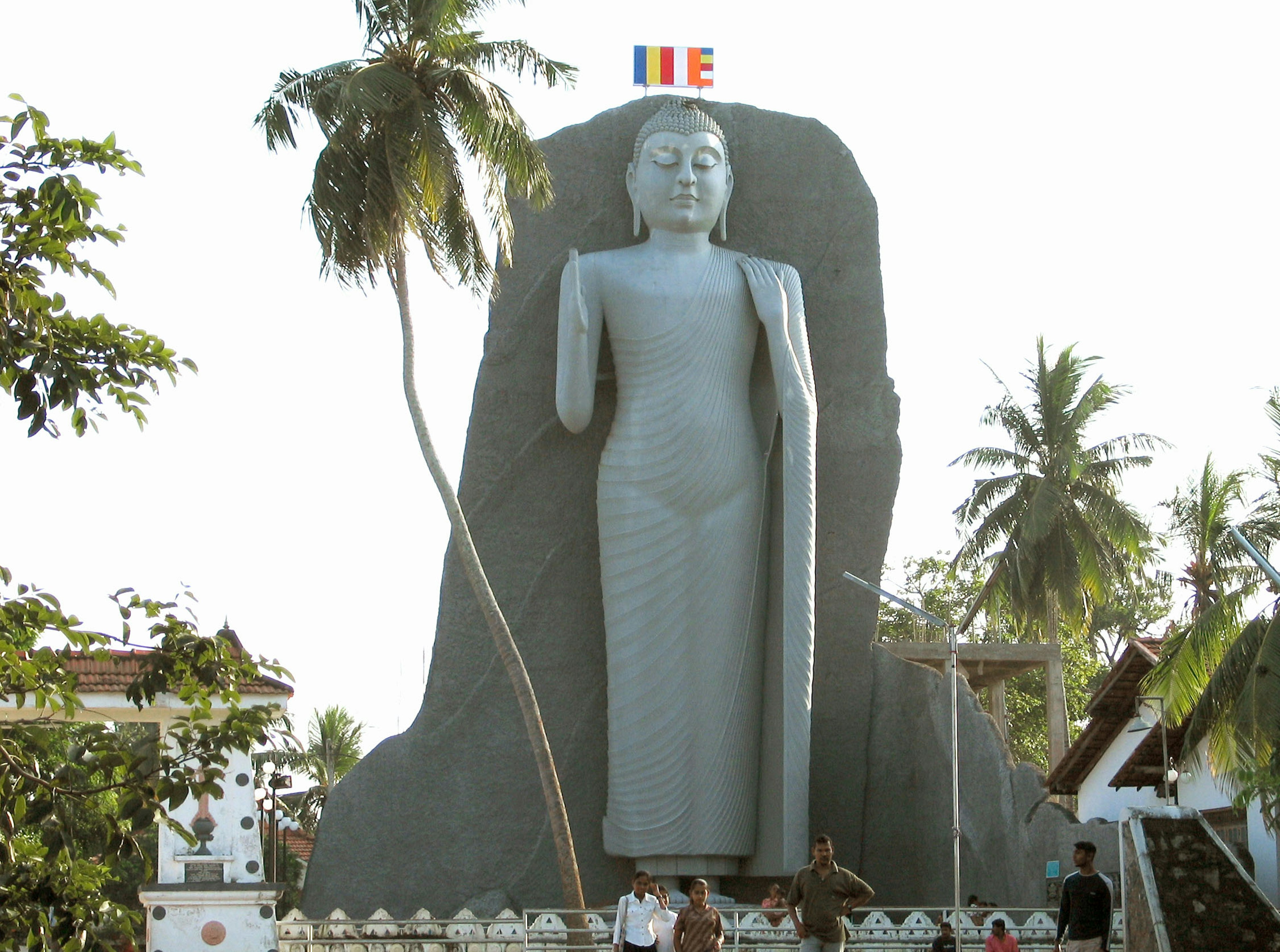 Sebuah patung Buddha besar berdiri dikelilingi pohon palem dan orang-orang