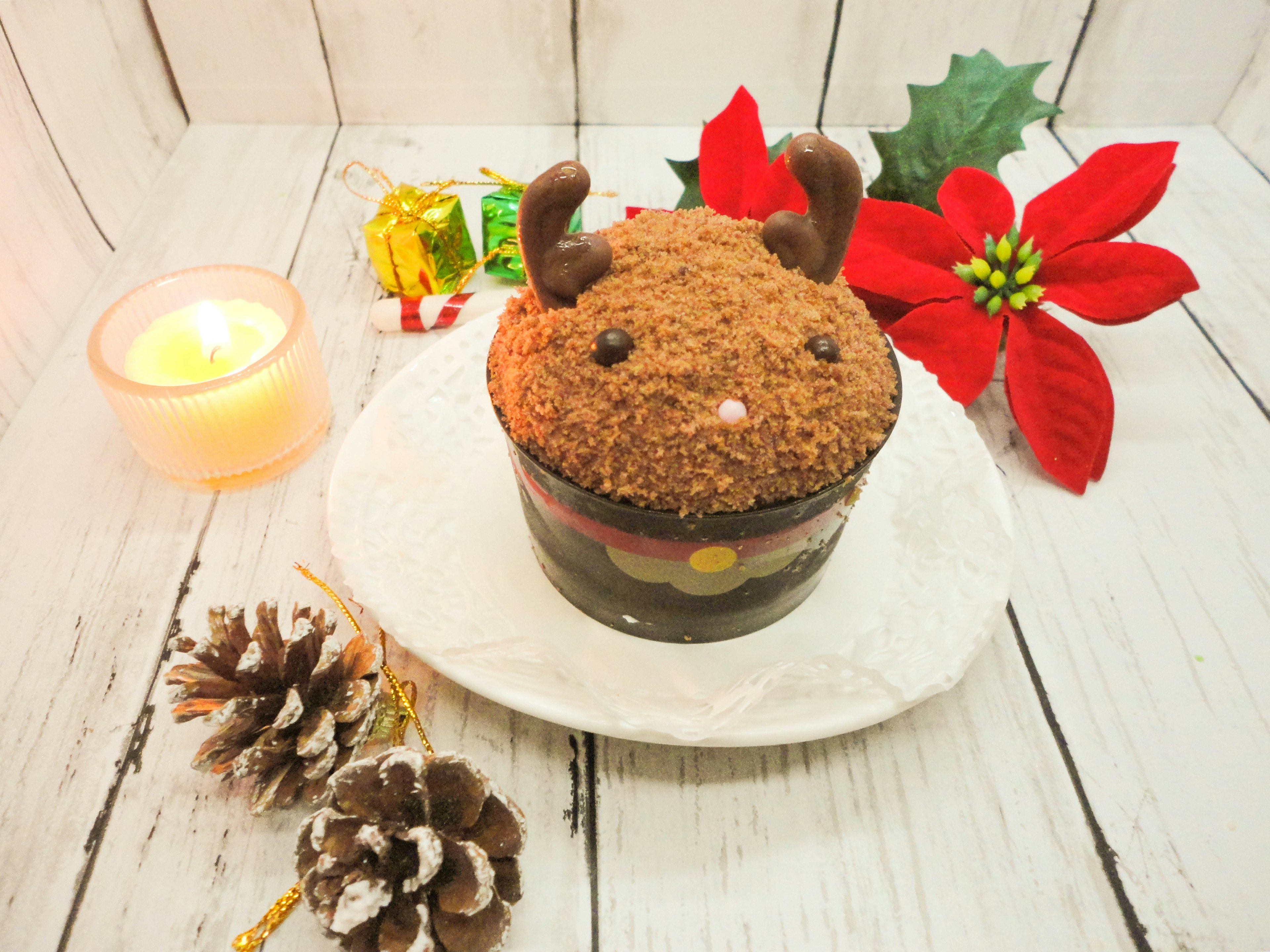 Una torta a forma di renna di Natale è posizionata su un piatto bianco circondata da una candela pigne e fiori di poinsettia