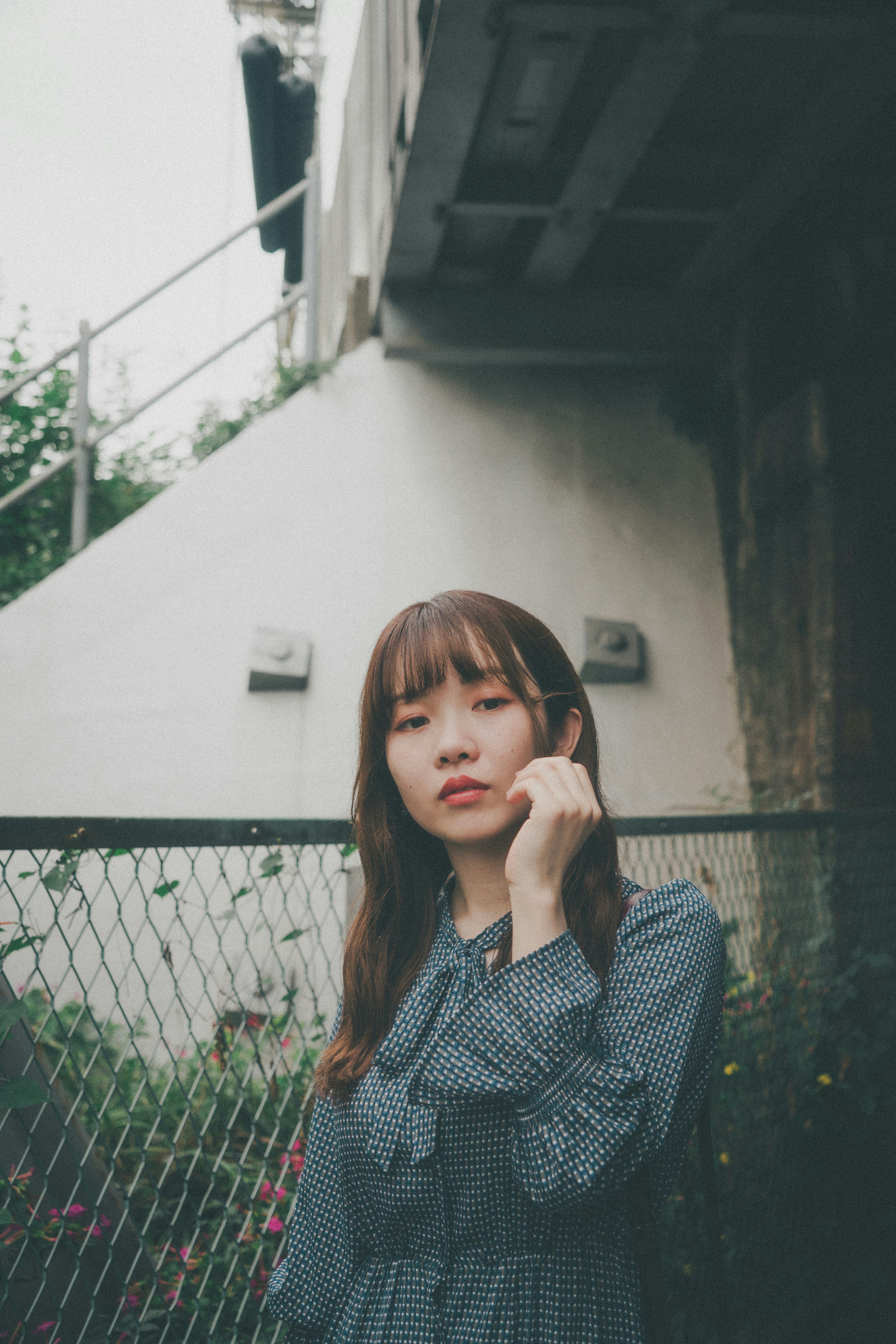 A woman in a gray dress poses outdoors with a fence and wall in the background