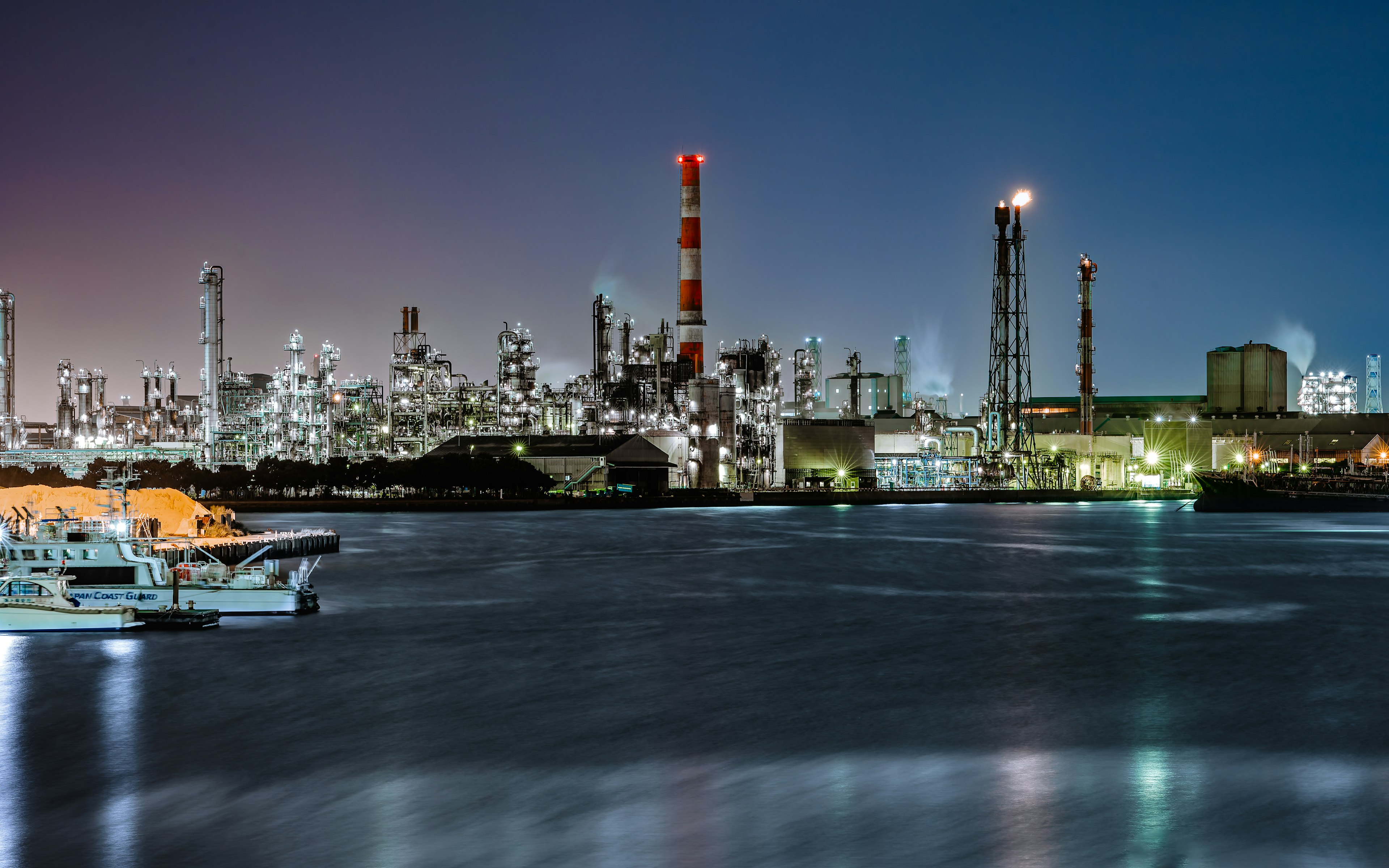 Nighttime industrial skyline featuring red smokestacks and a boat