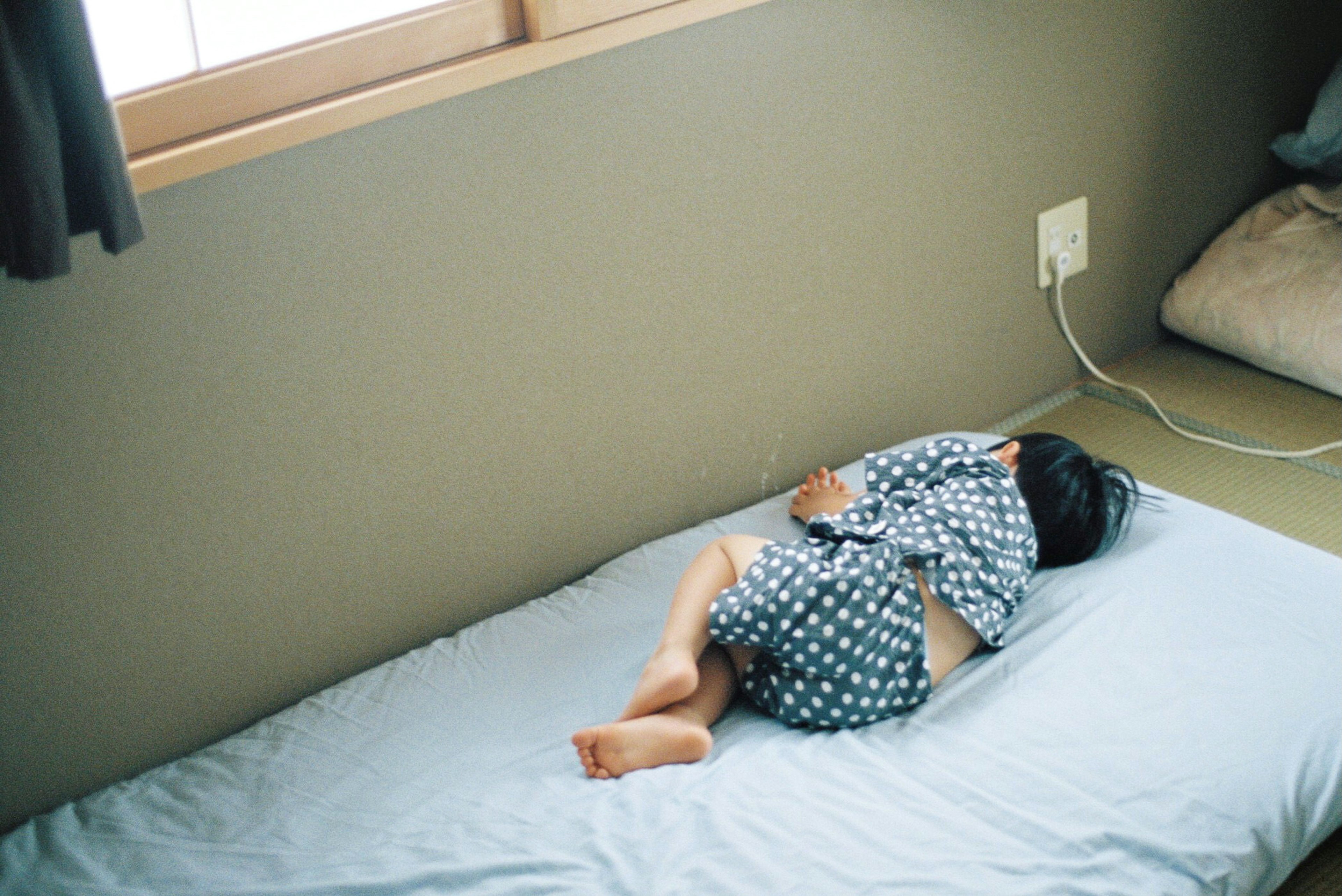 A child sleeping on a light blue bedspread wearing a blue polka dot pajama in a simple room