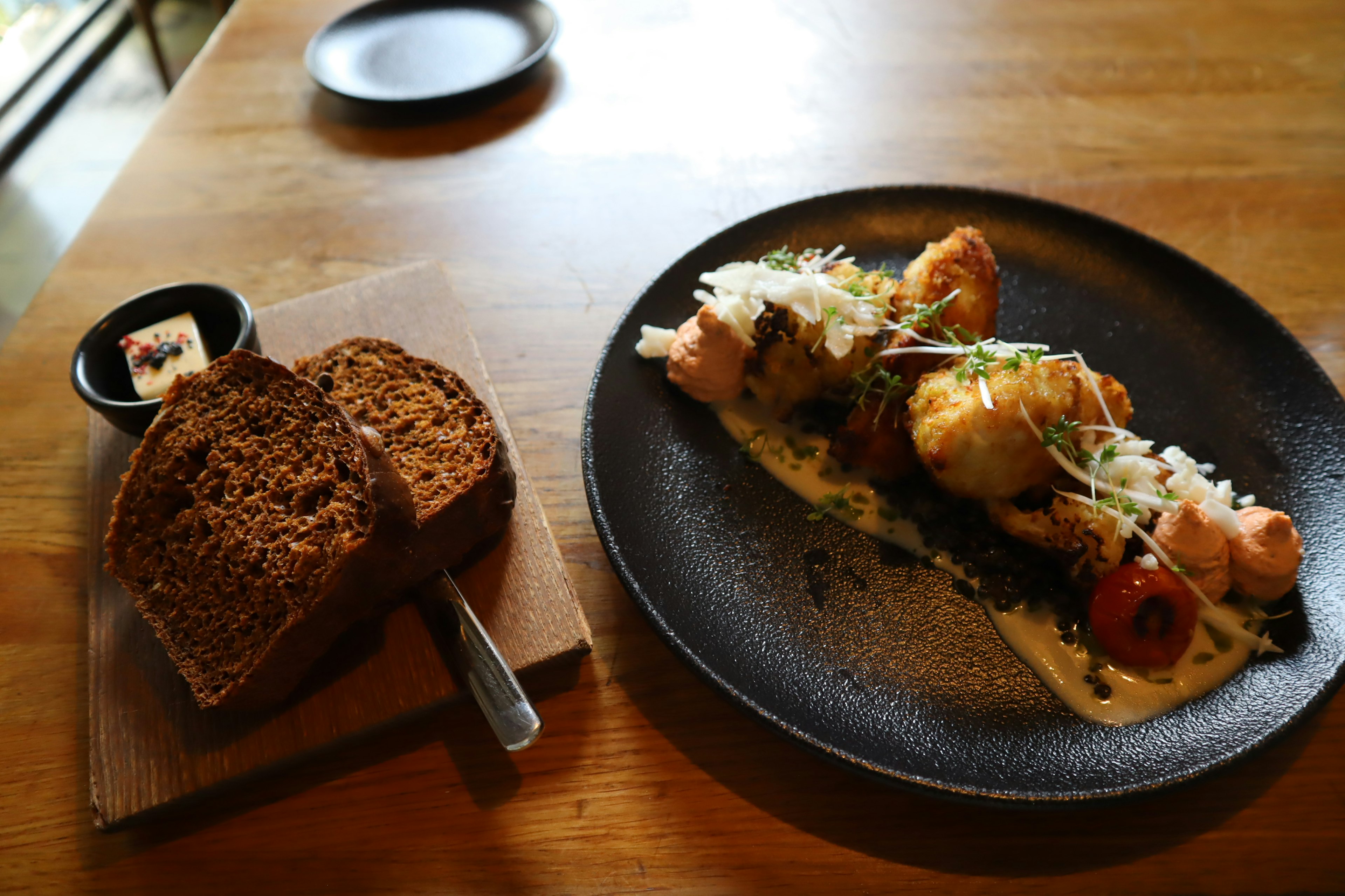 A beautifully plated fish dish on a black plate with sauce topped with tomatoes and herbs alongside a slice of dark bread