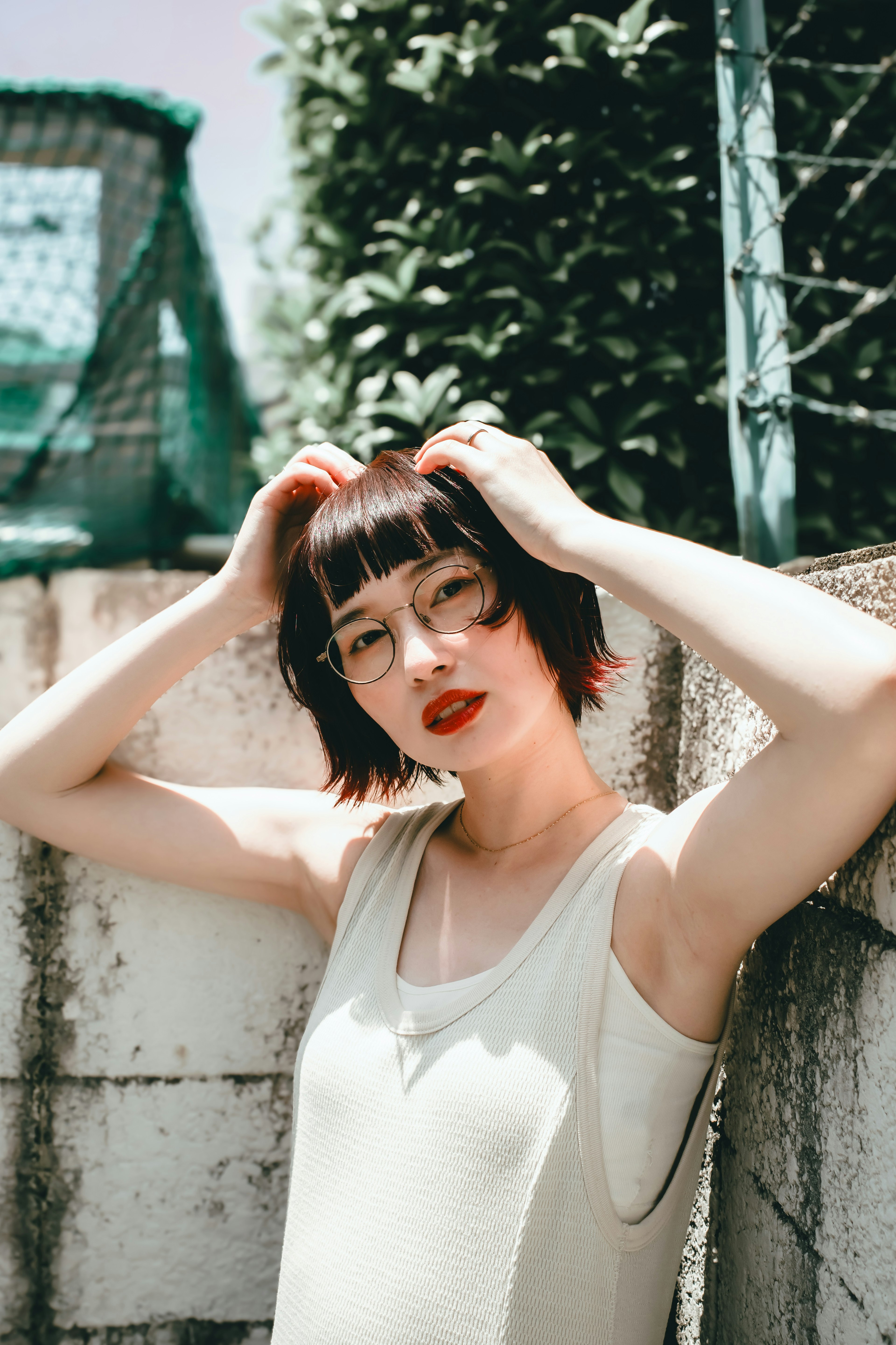 Una mujer con una camiseta blanca posando contra una pared texturizada con vegetación verde y una cerca de fondo