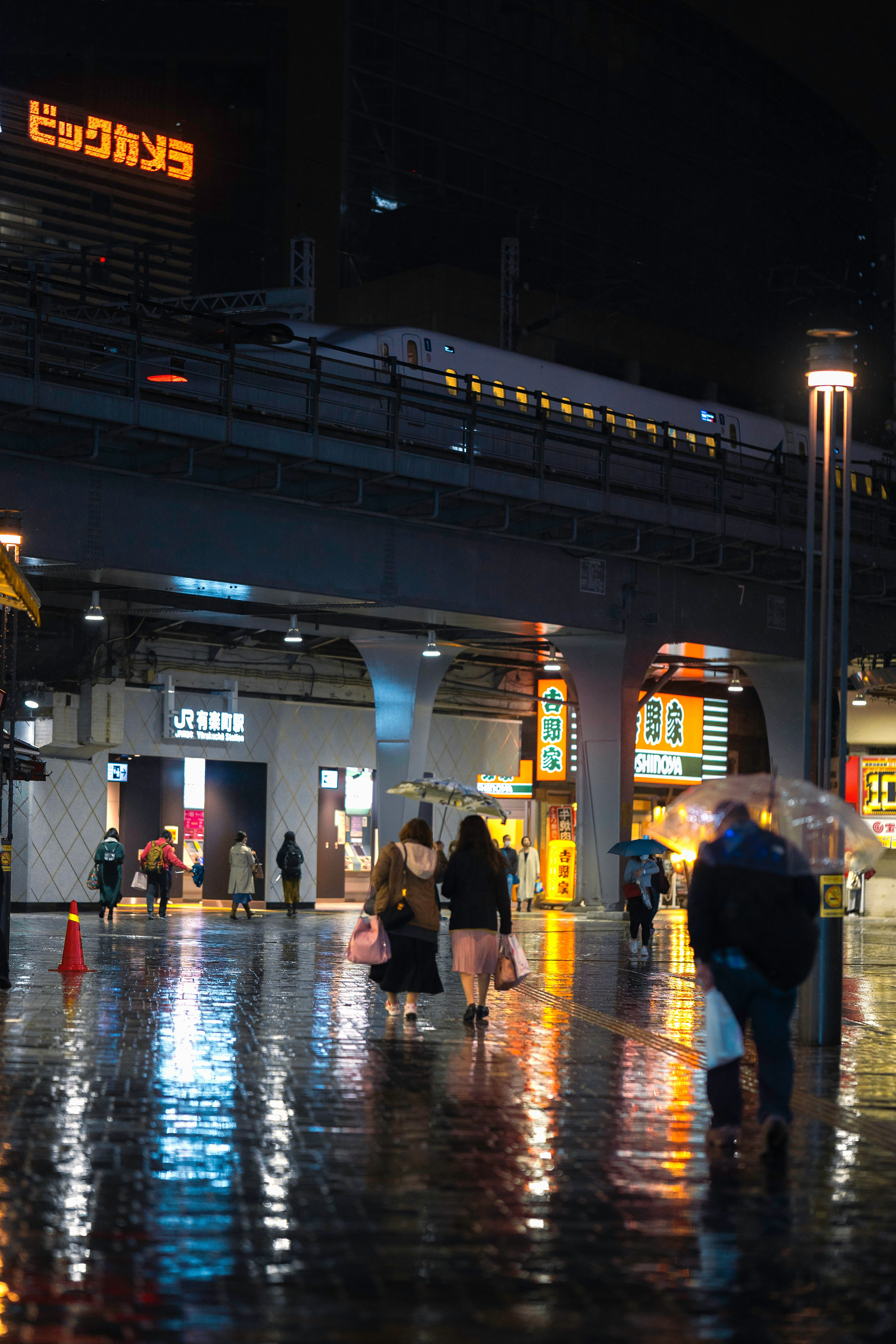 夜の駅で傘を持った人々が歩いている光景 鉄道と明るい看板が映る