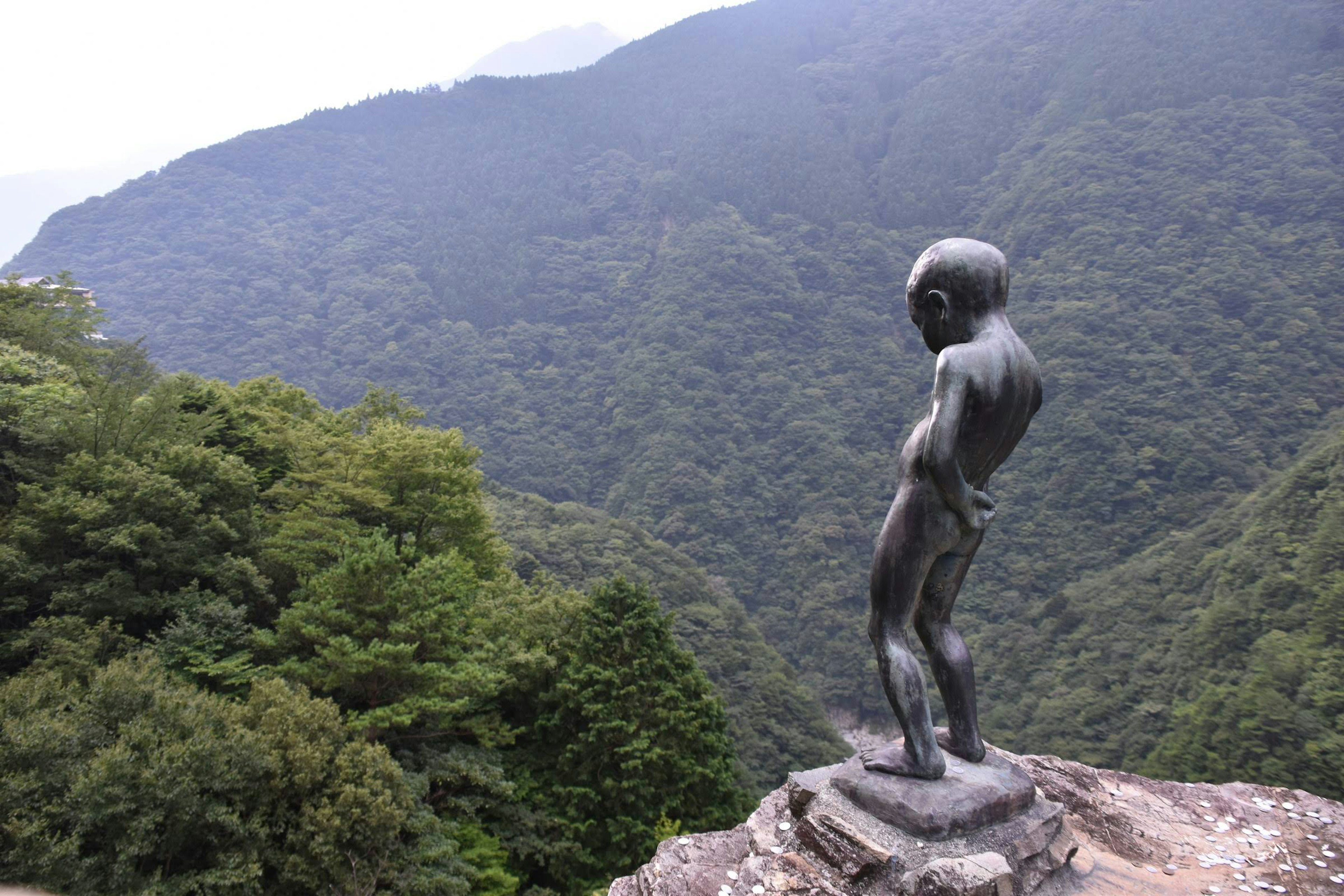 Kleine Bronzestatue, die auf einem Berg steht, umgeben von grünen Wäldern und Bergen