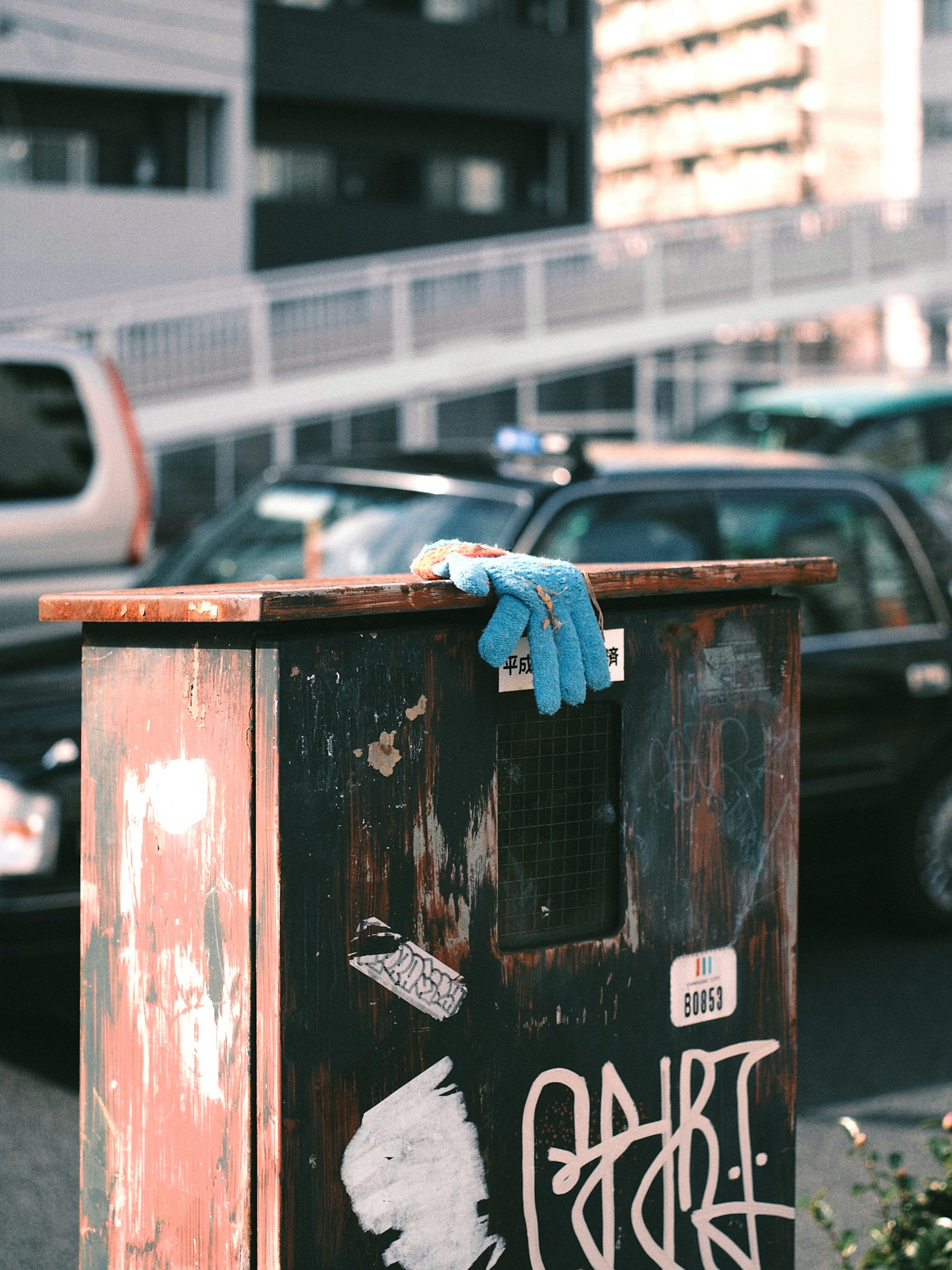 Un bote de basura con un guante azul encima y autos al fondo