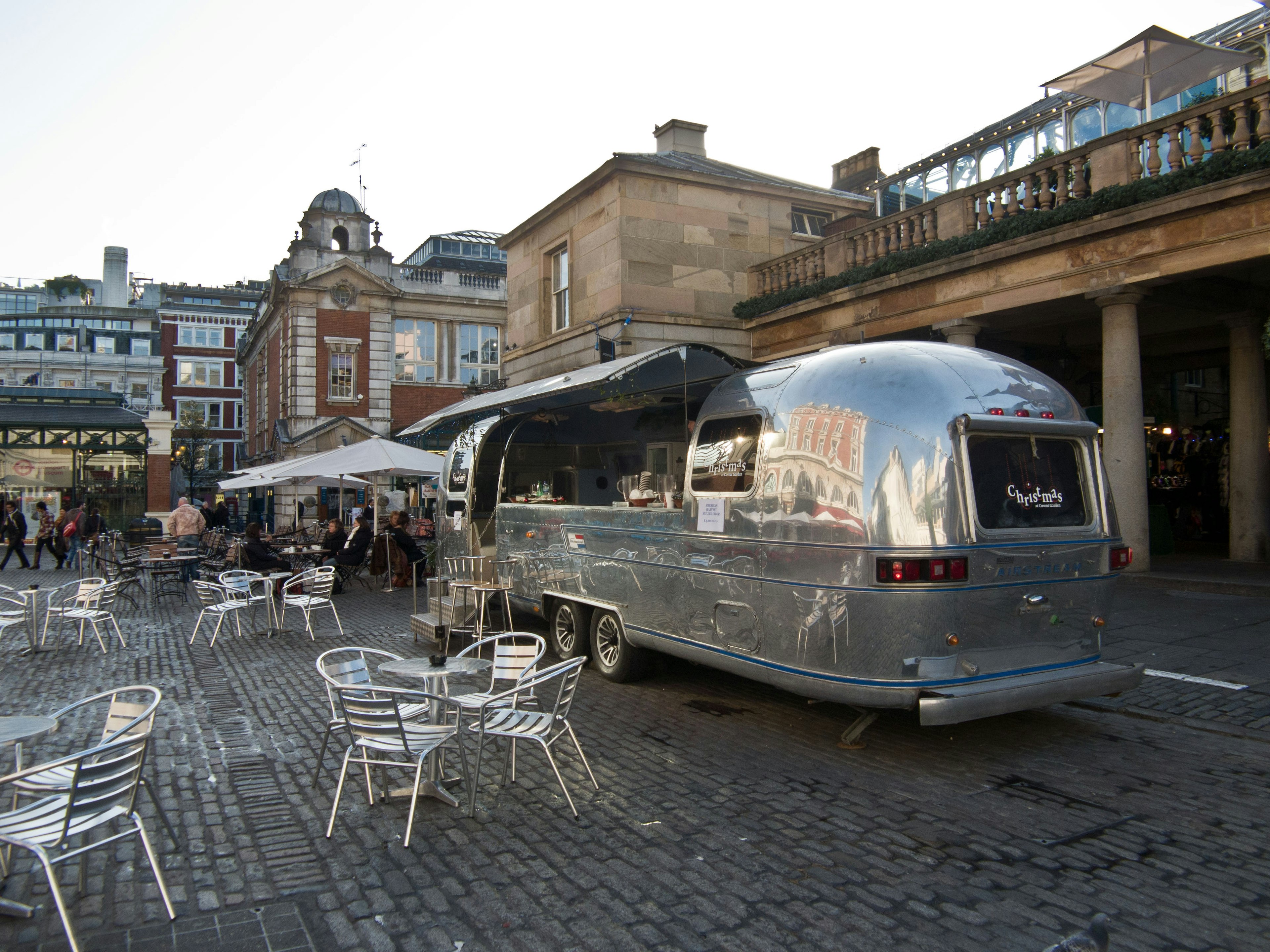 A silver food truck parked in a plaza with café tables and chairs arranged nearby