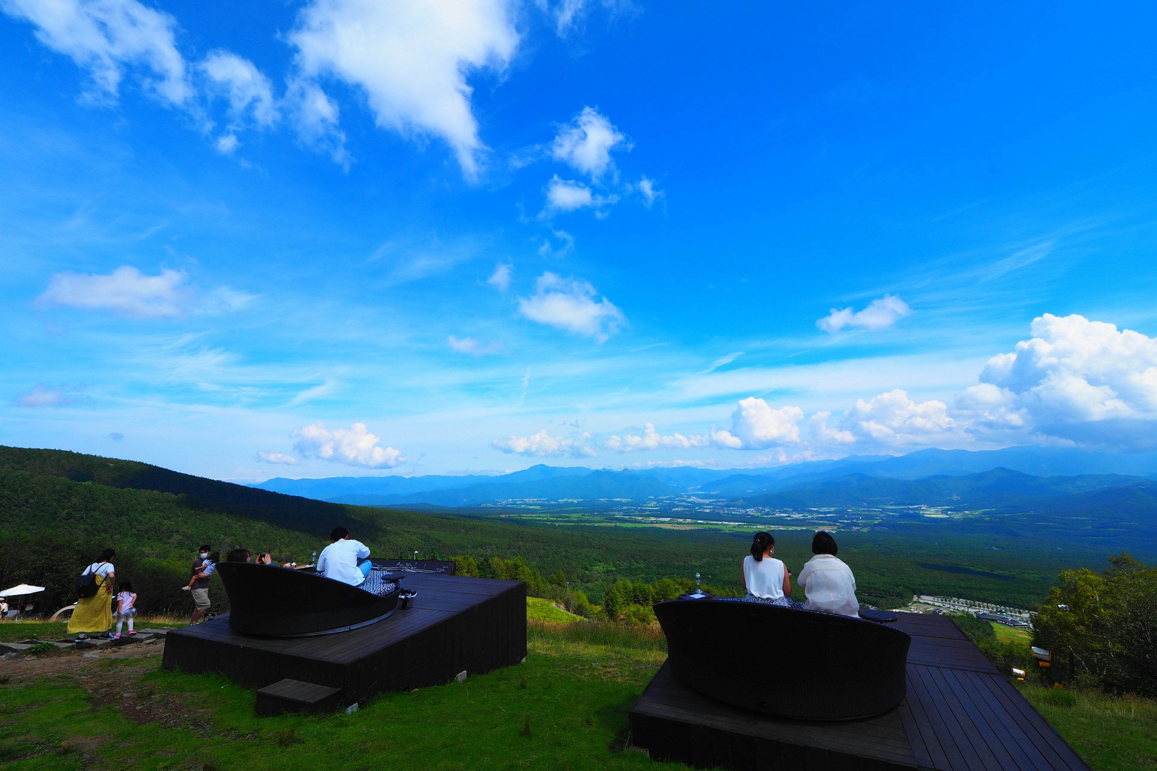 People sitting on benches overlooking a lush green valley under a bright blue sky