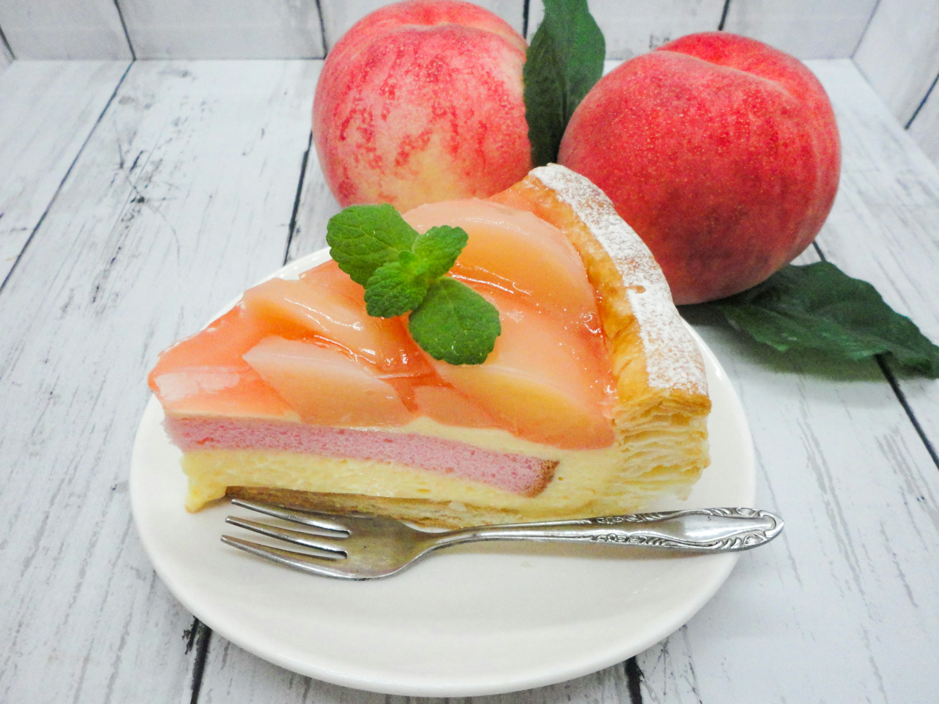 Una porción de pastel de durazno decorada con rodajas de durazno y hojas de menta junto a duraznos frescos