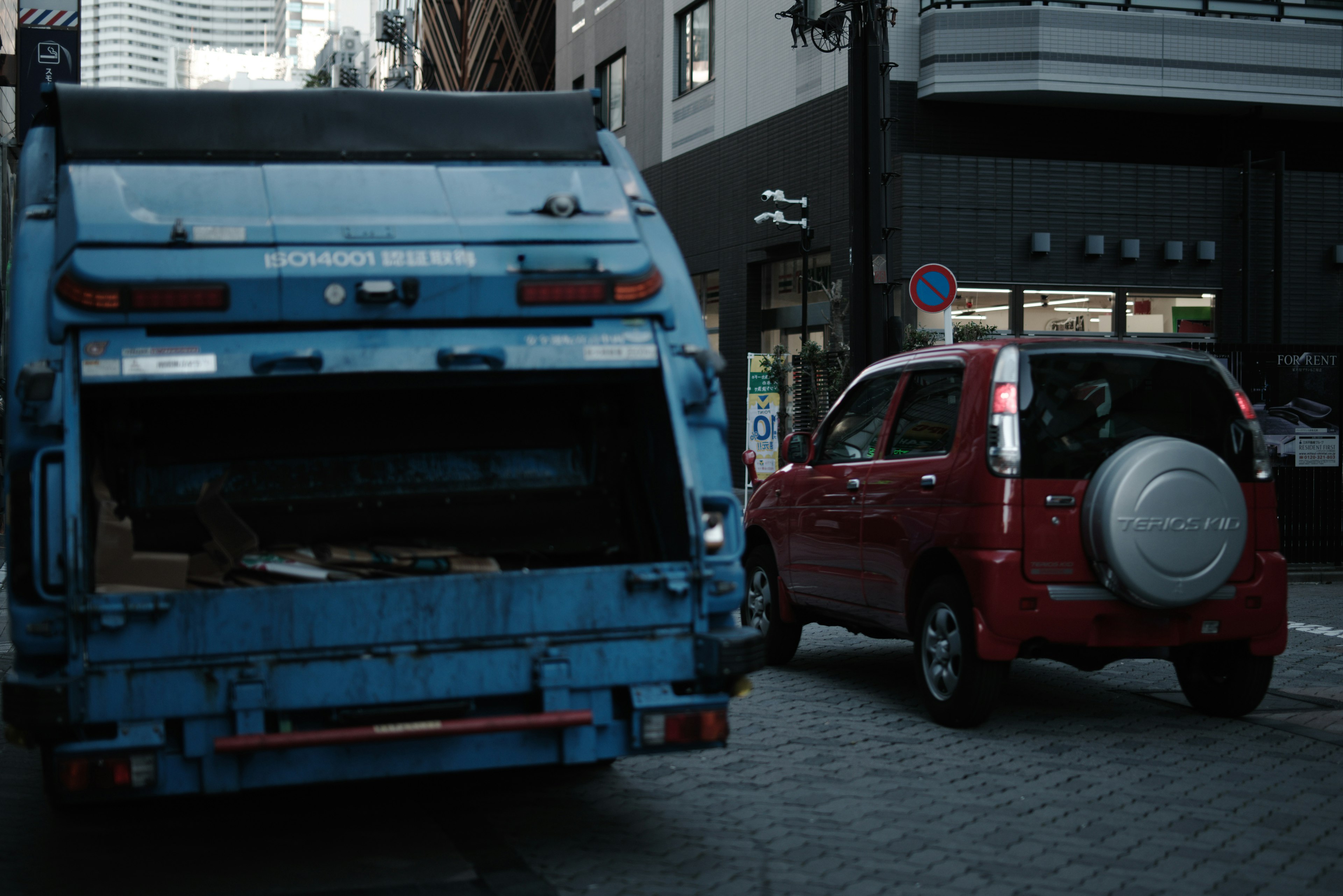 A blue garbage truck parked next to a red car on a city street