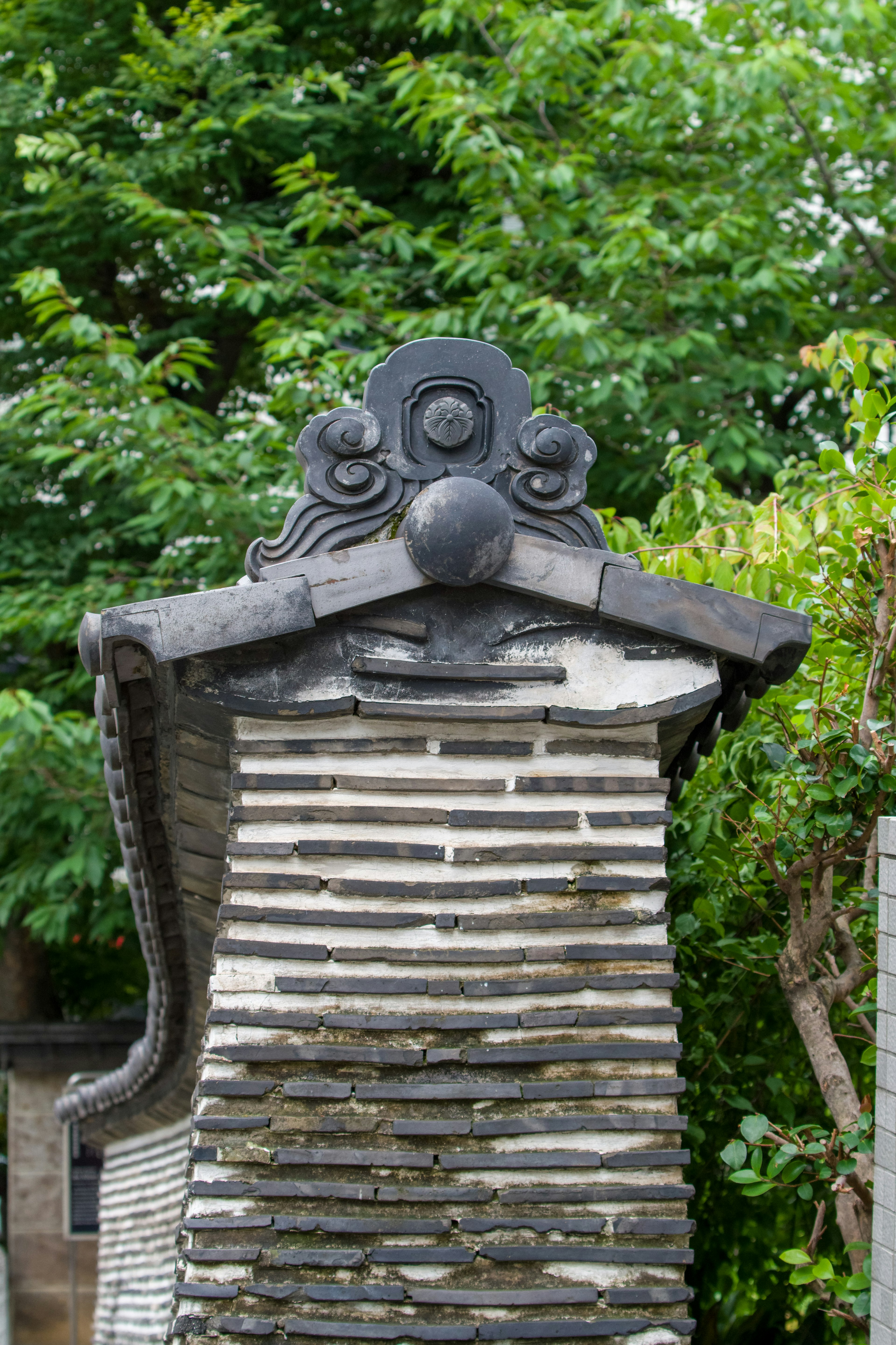 古い建物の屋根の一部が見え、黒い瓦と石の壁が特徴的で、背景には緑の木々がある