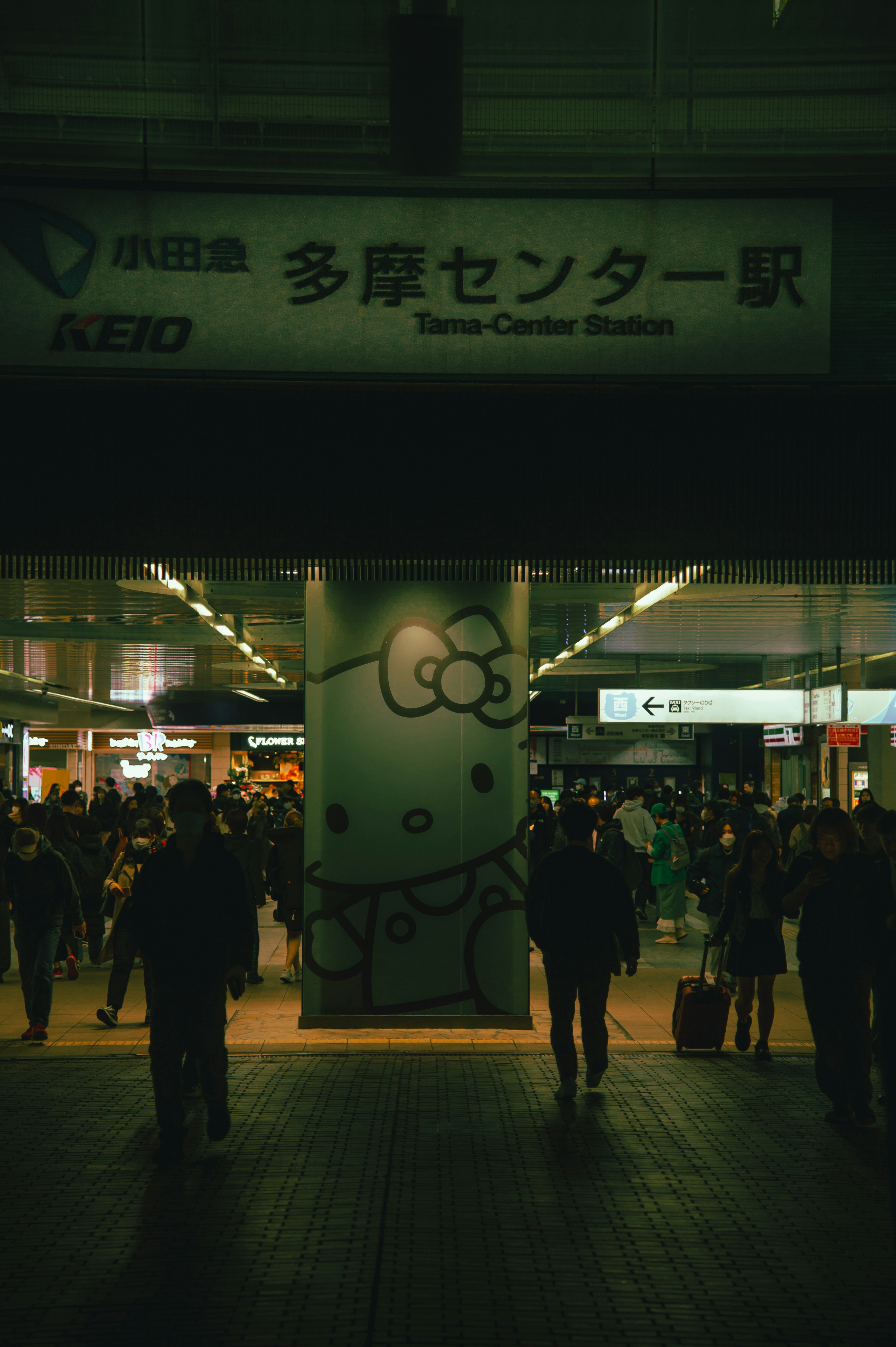 Panneau de la station Tama Center avec une illustration de Hello Kitty et des gens qui passent