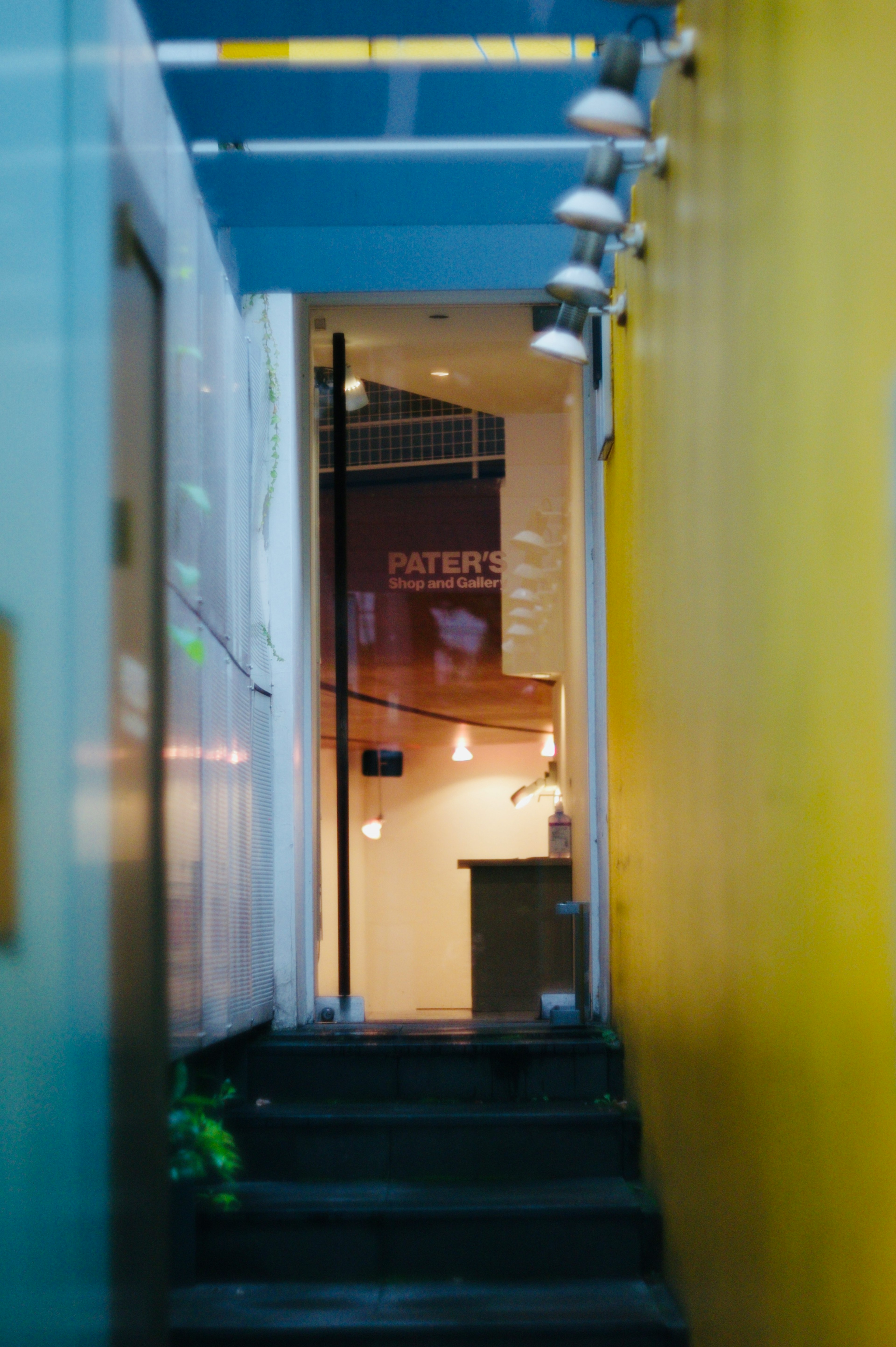 Narrow alleyway flanked by bright yellow walls leading to a warmly lit room