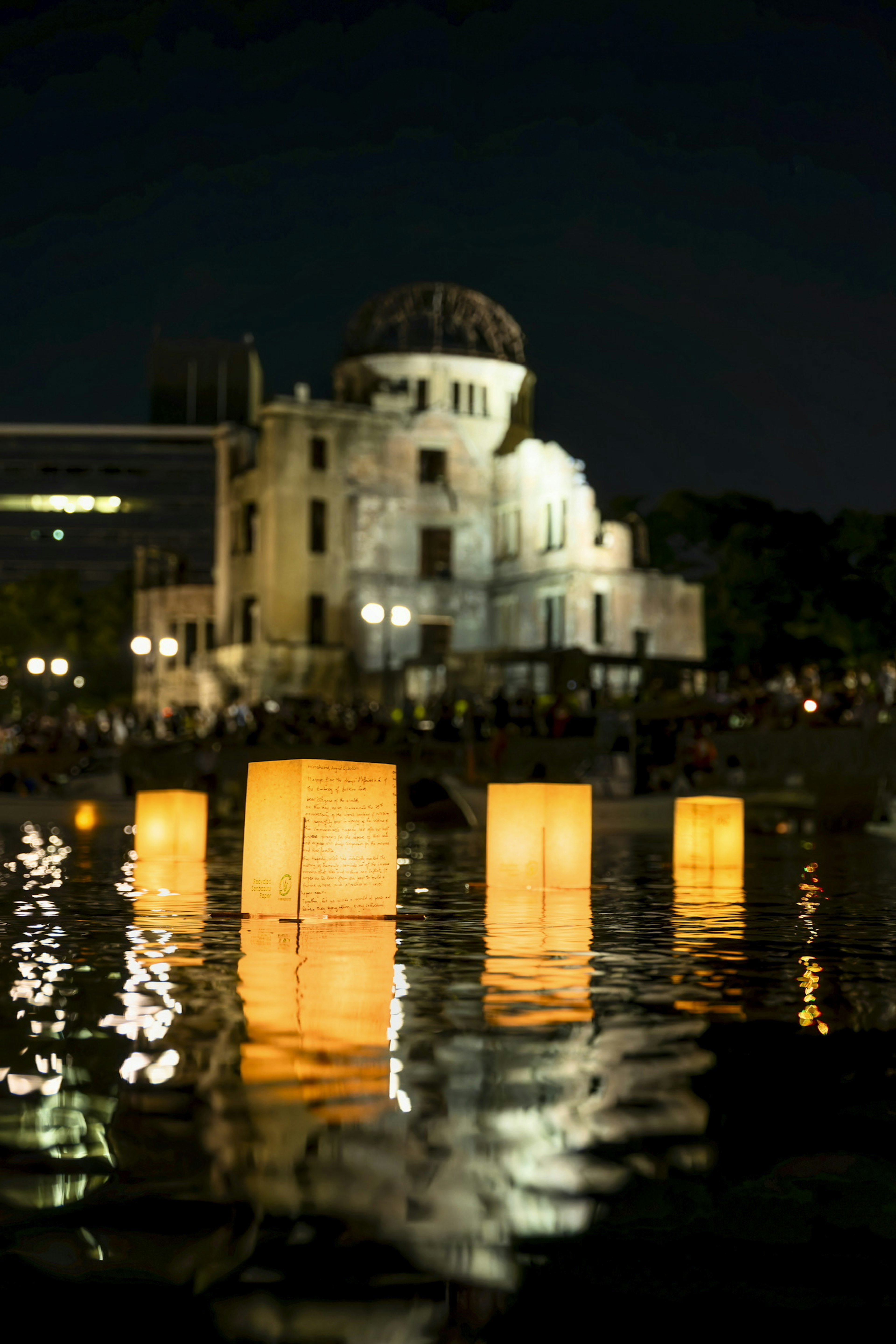 Đèn lồng sáng rực nổi trên mặt nước tại Công viên Tưởng niệm Hòa bình Hiroshima với Cột mốc Bom Nguyên tử ở phía sau