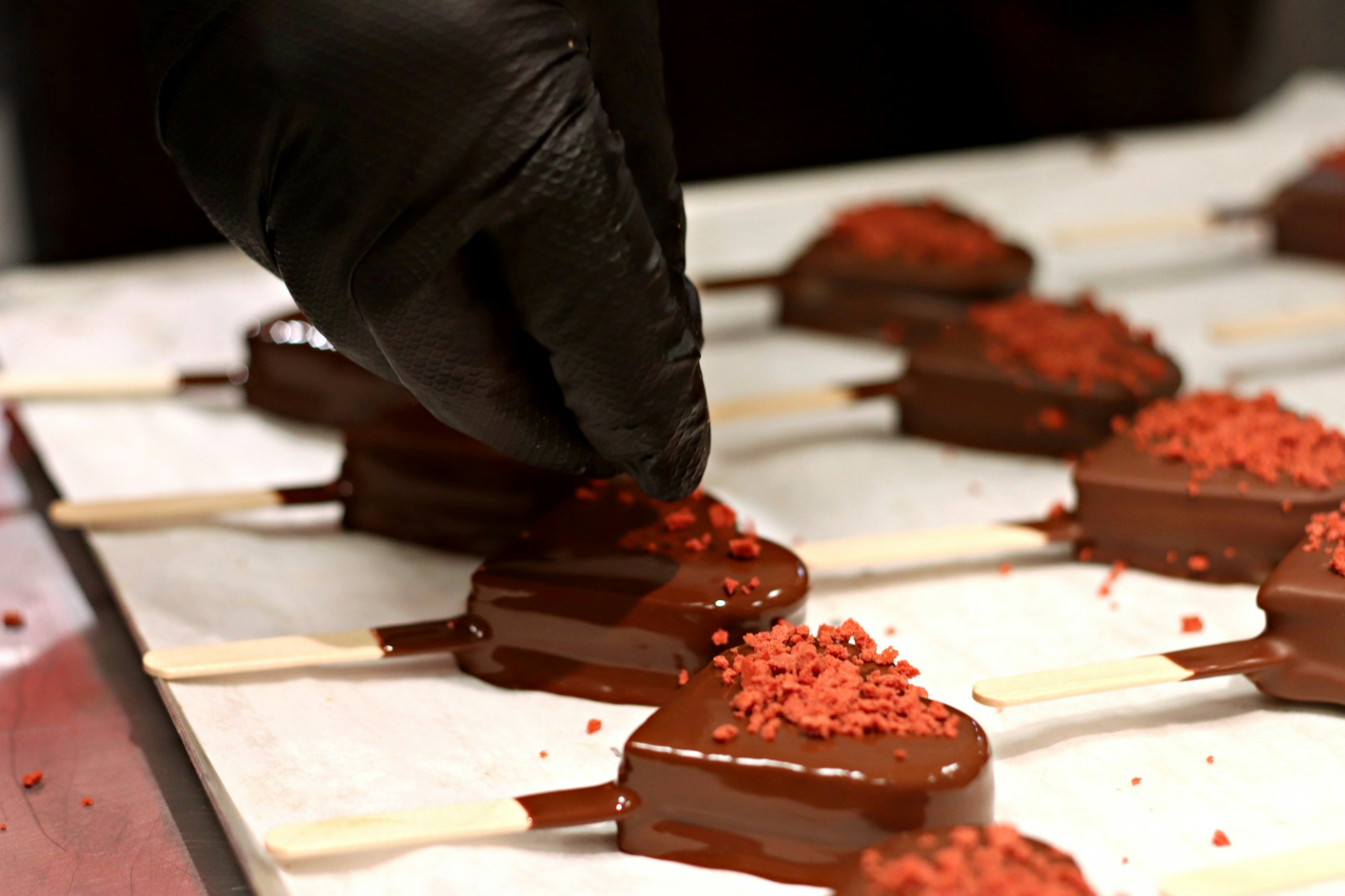 Una mano añadiendo chispas rojas a paletas de hielo en forma de corazón cubiertas de chocolate