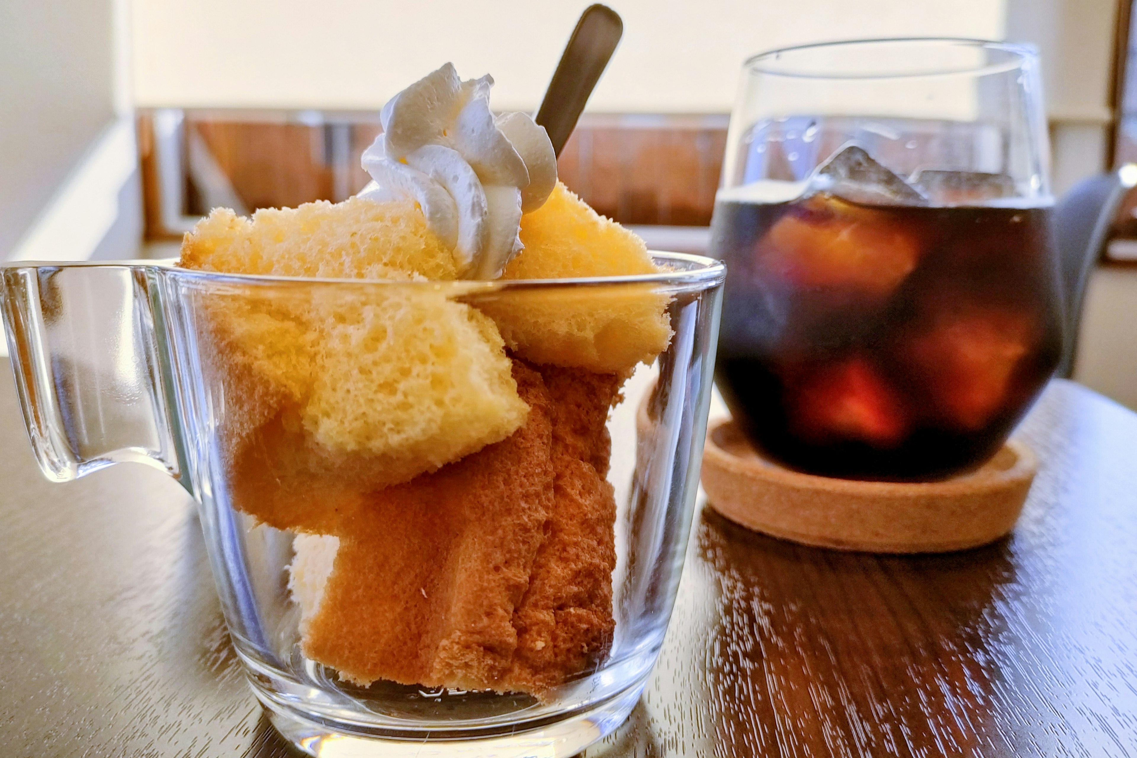 Dessert in einer klaren Tasse mit Schwammkuchen und Schlagsahne neben einem Glas Eiskaffee