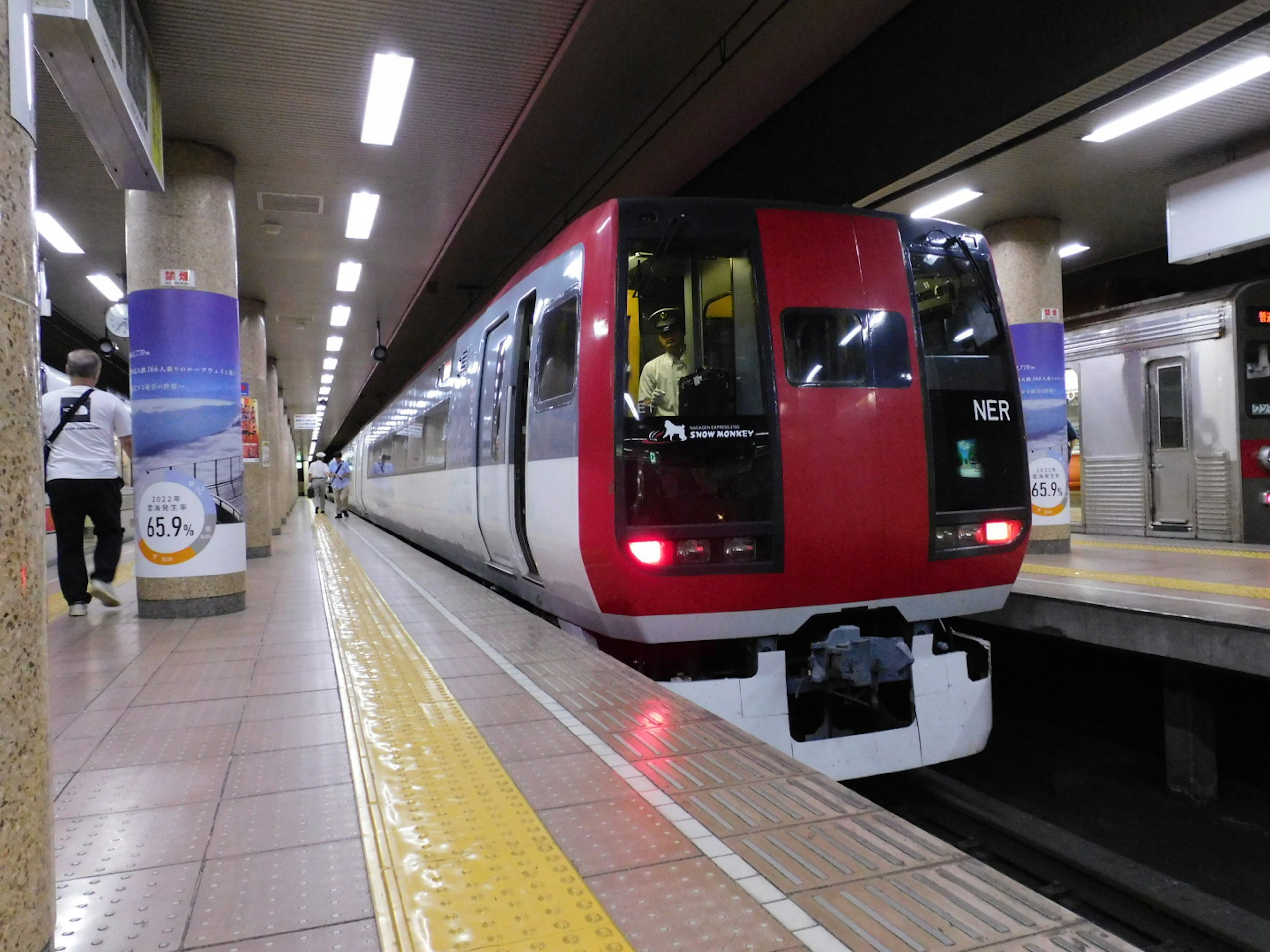 Train rouge arrêté à une plateforme de gare avec des panneaux visibles